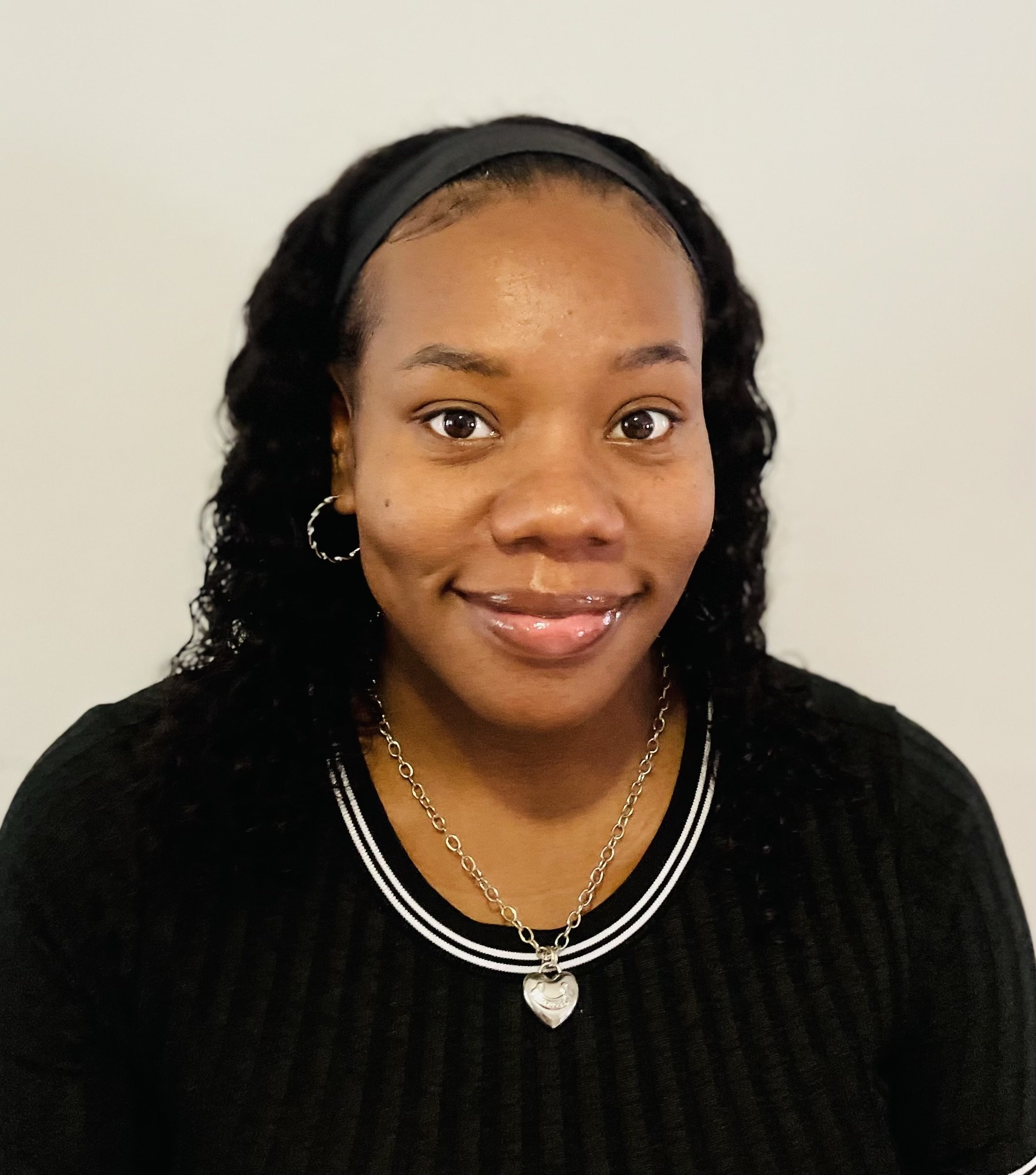 Portrait of a smiling woman with curly hair, wearing a black headband, hoop earrings, a silver necklace with a heart pendant, and a black top with white trim, standing against a plain light-colored background.