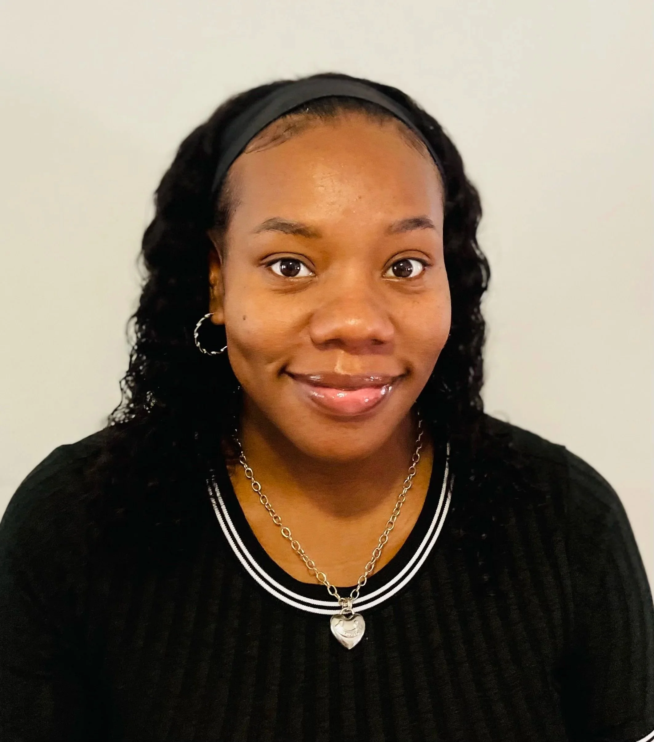 A smiling woman with dark, curly hair wearing a black headband, hoop earrings, and a necklace with a heart-shaped pendant. She is dressed in a black top with white stripes around the neckline, standing against a plain light background.
