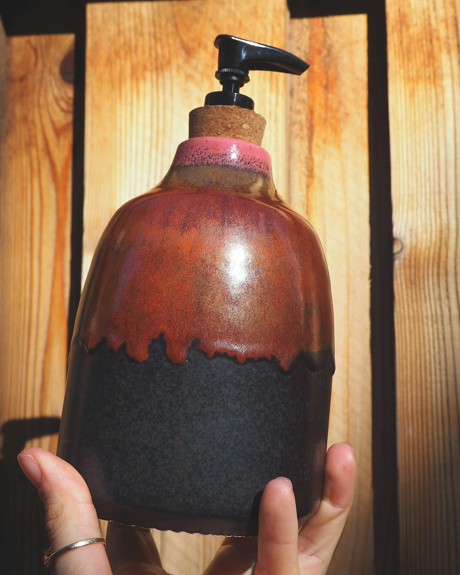 Hand holding the red and black ceramic soap dispenser against a sunlit fence
