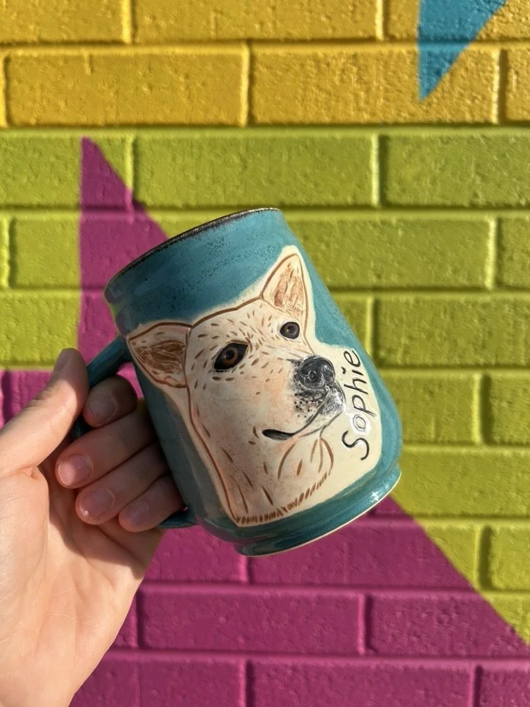 Teal mug with a carved cream dog portrait named Sophie, held against a colorful wall