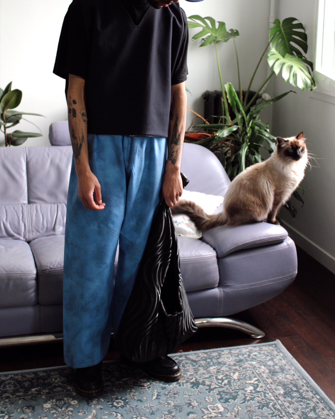 Personnes dans un salon avec un chat siamois assis sur un canapé en cuir gris, entouré de plantes vertes, près d'une fenêtre.