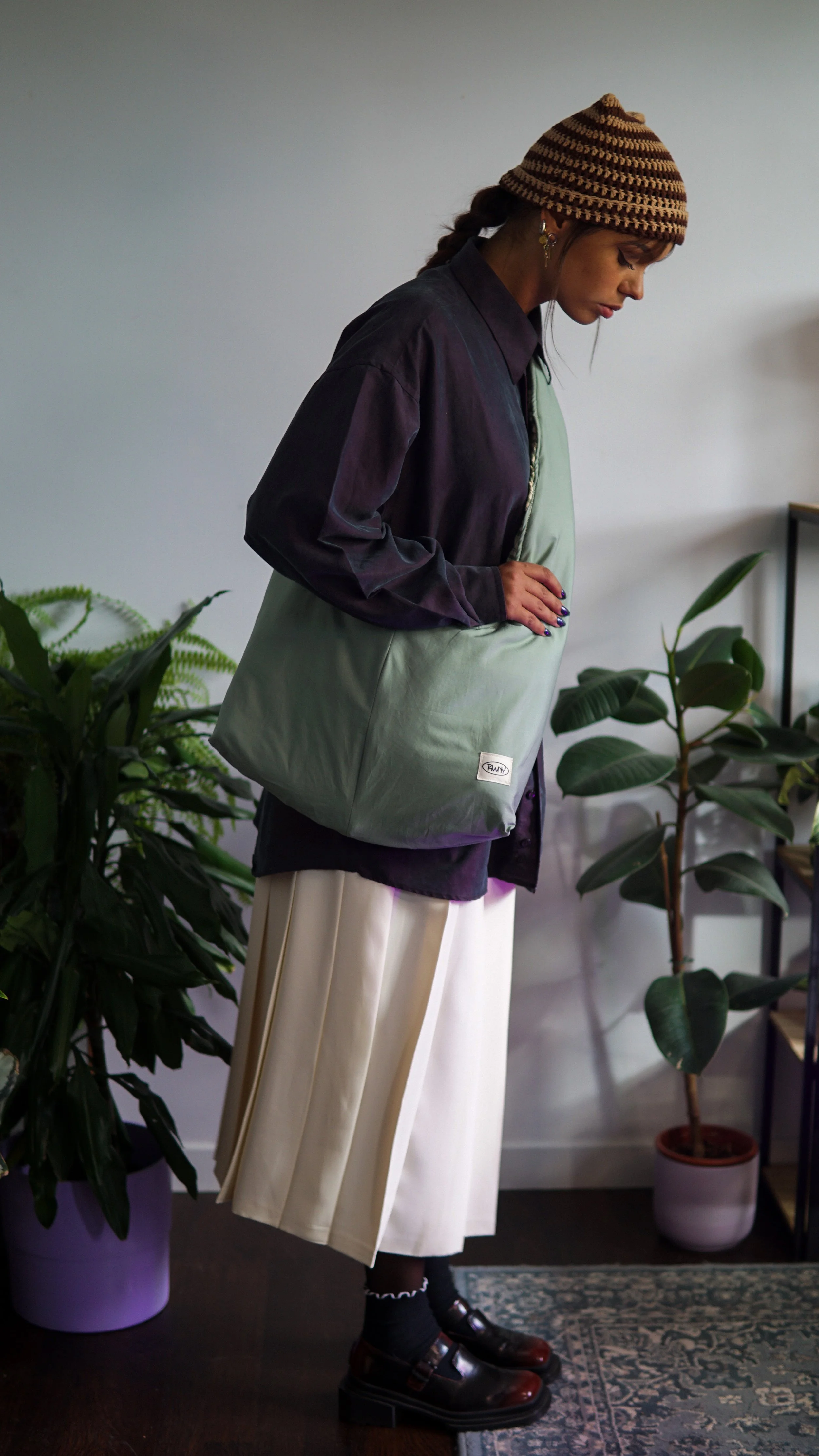 Une femme portant un bonnet en tricot, une veste à motifs foncés, un sac vert, une jupe plissée blanche, des chaussettes noires et des chaussures en cuir marron, se tient à côté de plantes d'intérieur dans une pièce.