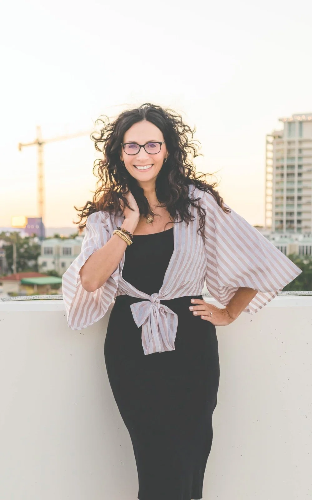 Woman with dark curly hair and glasses smiling, standing outdoors on a balcony with buildings in the background during sunset.