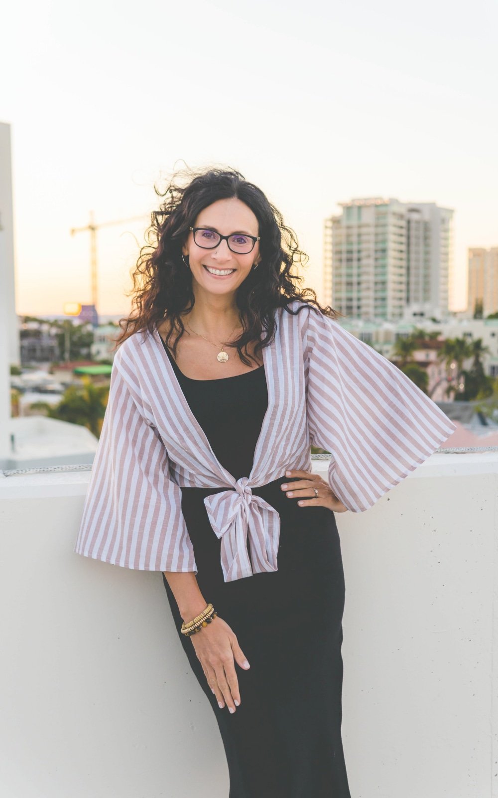 Gail Gerbig, therapist at Seaside Mental Wellness, a woman with curly hair, glasses, and wearing a black dress, smiles while on a rooftop in downtown Sarasota at sunset.