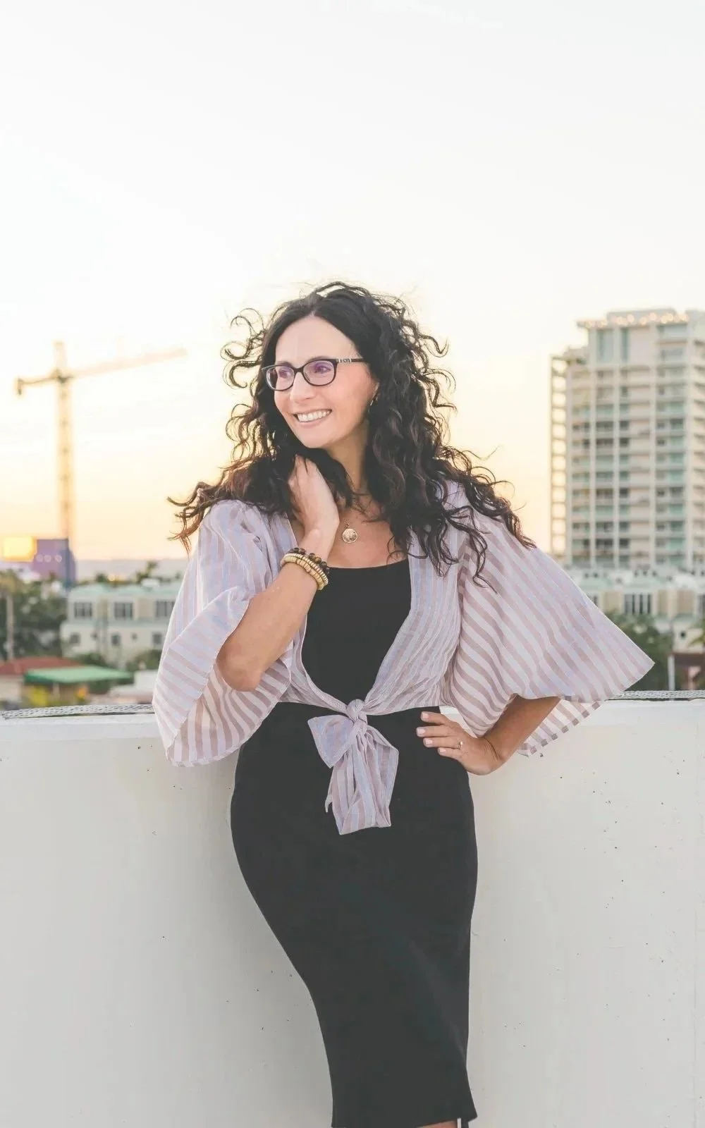 Gail Gerbig, therapist at Seaside Mental Wellness, a woman with curly hair, glasses, and wearing a black dress, smiles while on a rooftop in downtown Sarasota at sunset.