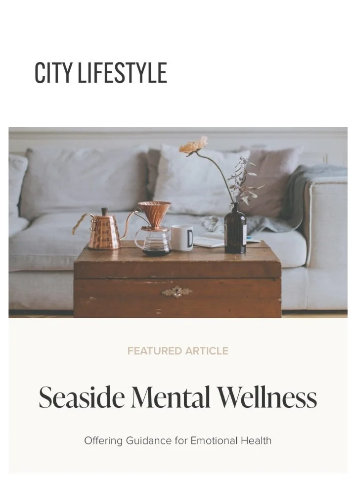 A cozy living room with a beige sofa, a wooden coffee table with a teapot, a glass pour-over coffee maker, a white mug, a black vase with a flower, and a book, with a white wall in the background.