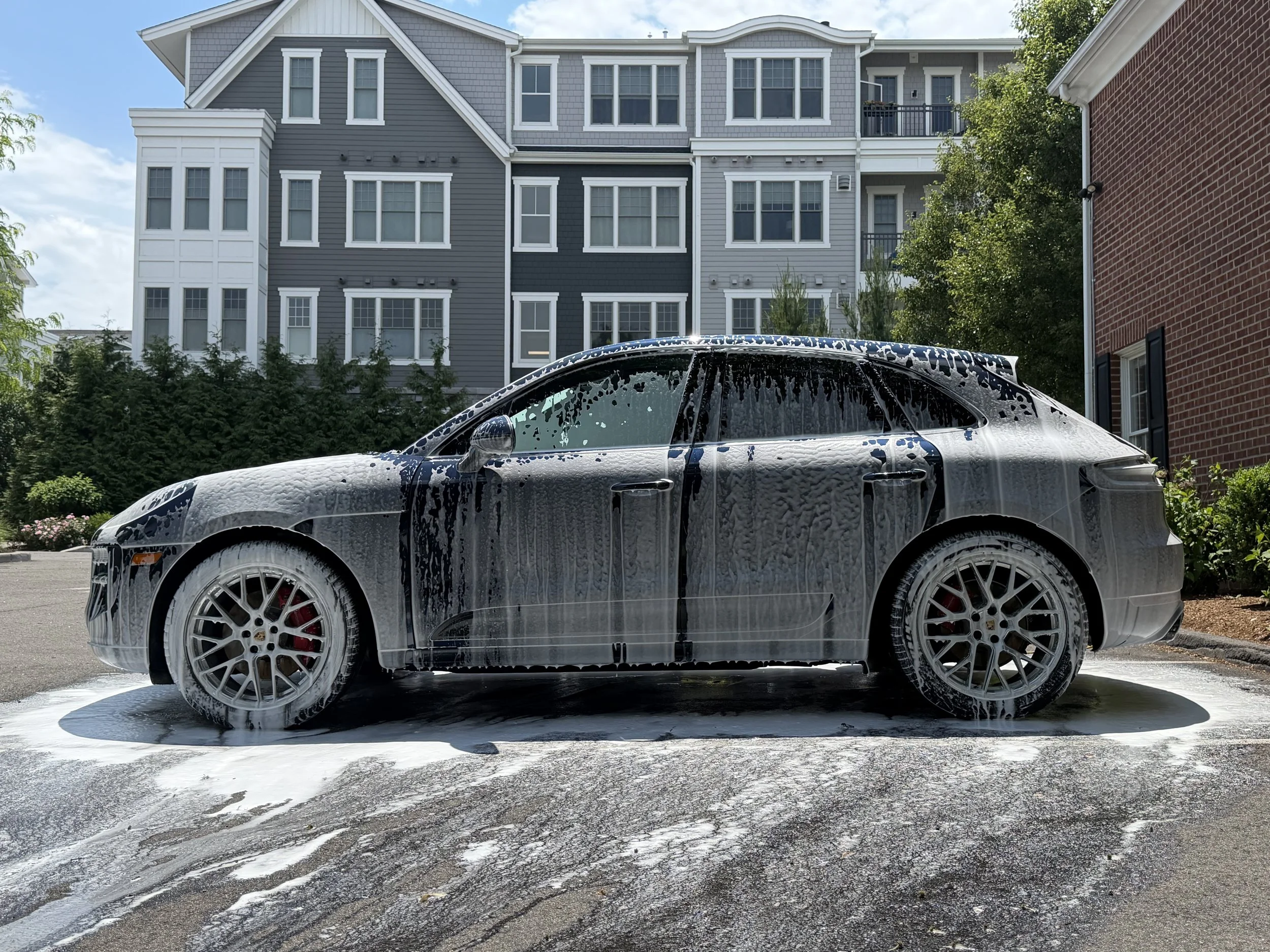 Car covered in soap suds during a car wash, with a residential building and trees in the background.