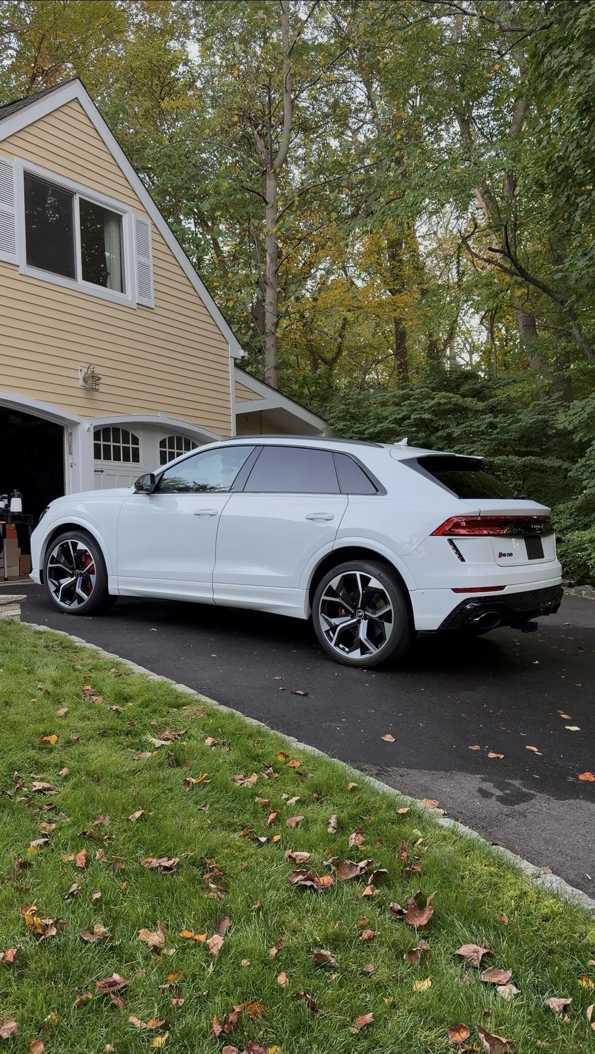 A white Audi SUV parked in a driveway in front of a beige house with a garage door, green trees with autumn leaves, and a small lawn with fallen leaves.
