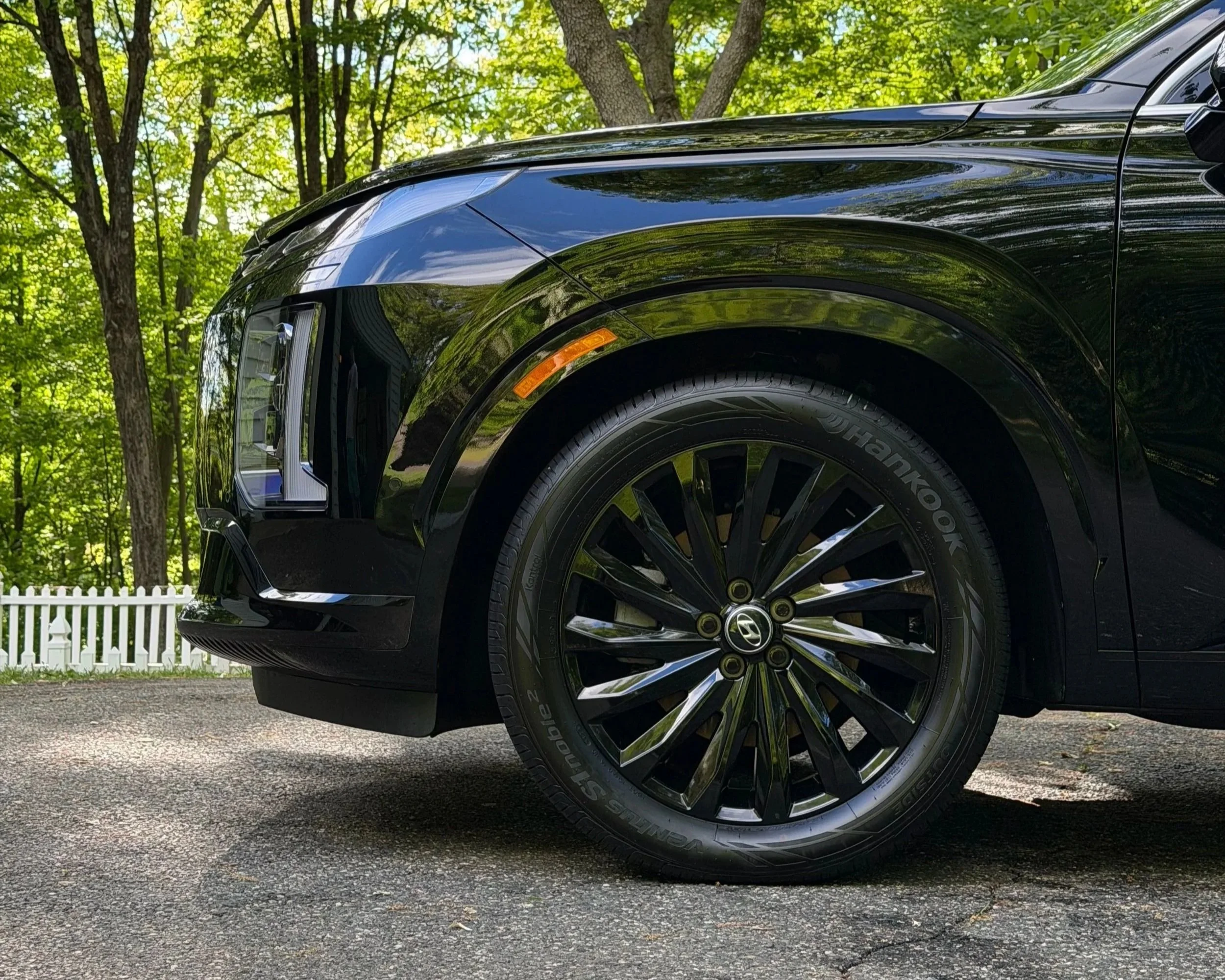 Close-up of the front side of a shiny black car, showing the headlight, tire, and part of the front bumper, with a background of green trees and a white picket fence.