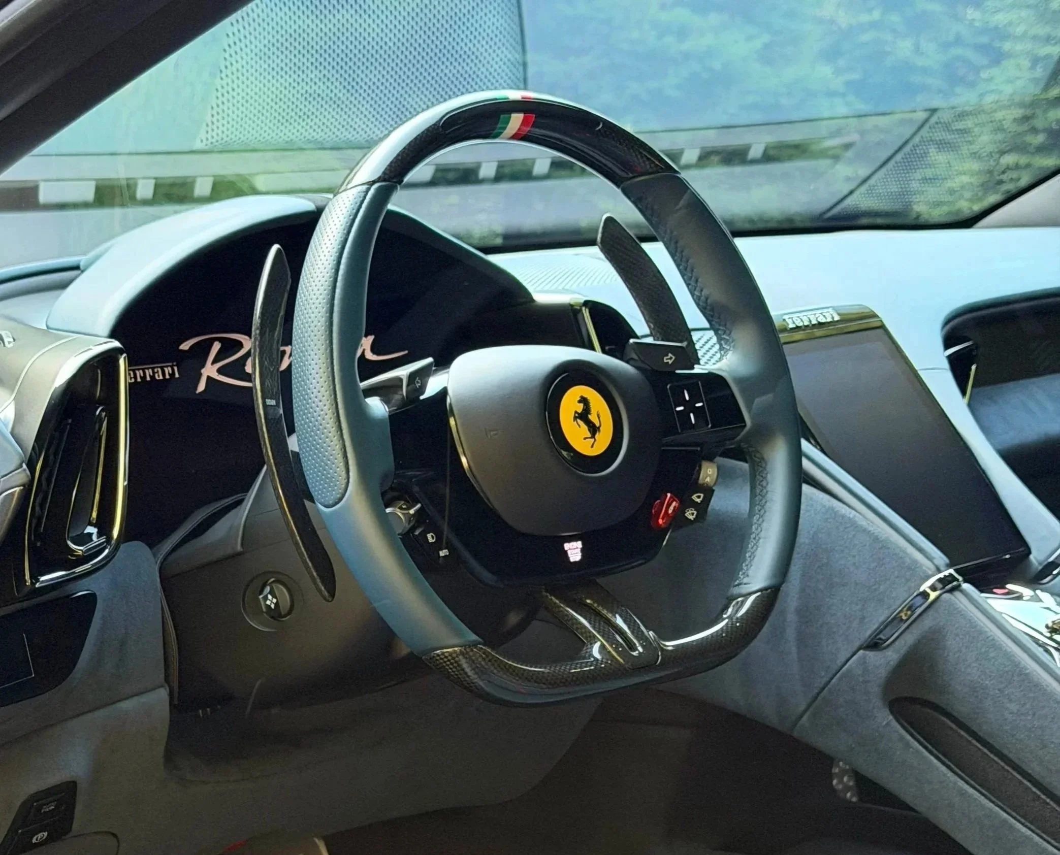 Interior of a Ferrari car steering wheel with a yellow Ferrari logo in the center, dashboard, and part of a digital display, with the background showing a blurred view of the road.