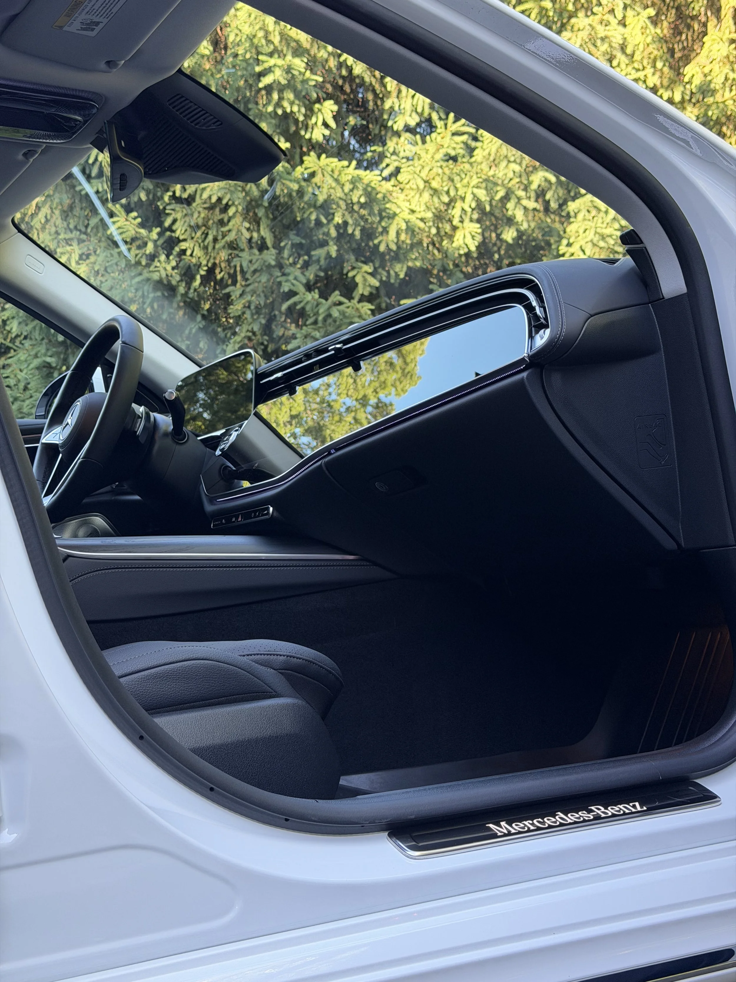 Interior view of a white Mercedes-Benz vehicle showing the dashboard, steering wheel, passenger seat, and a large sunroof with a scenic outdoor view of green trees.