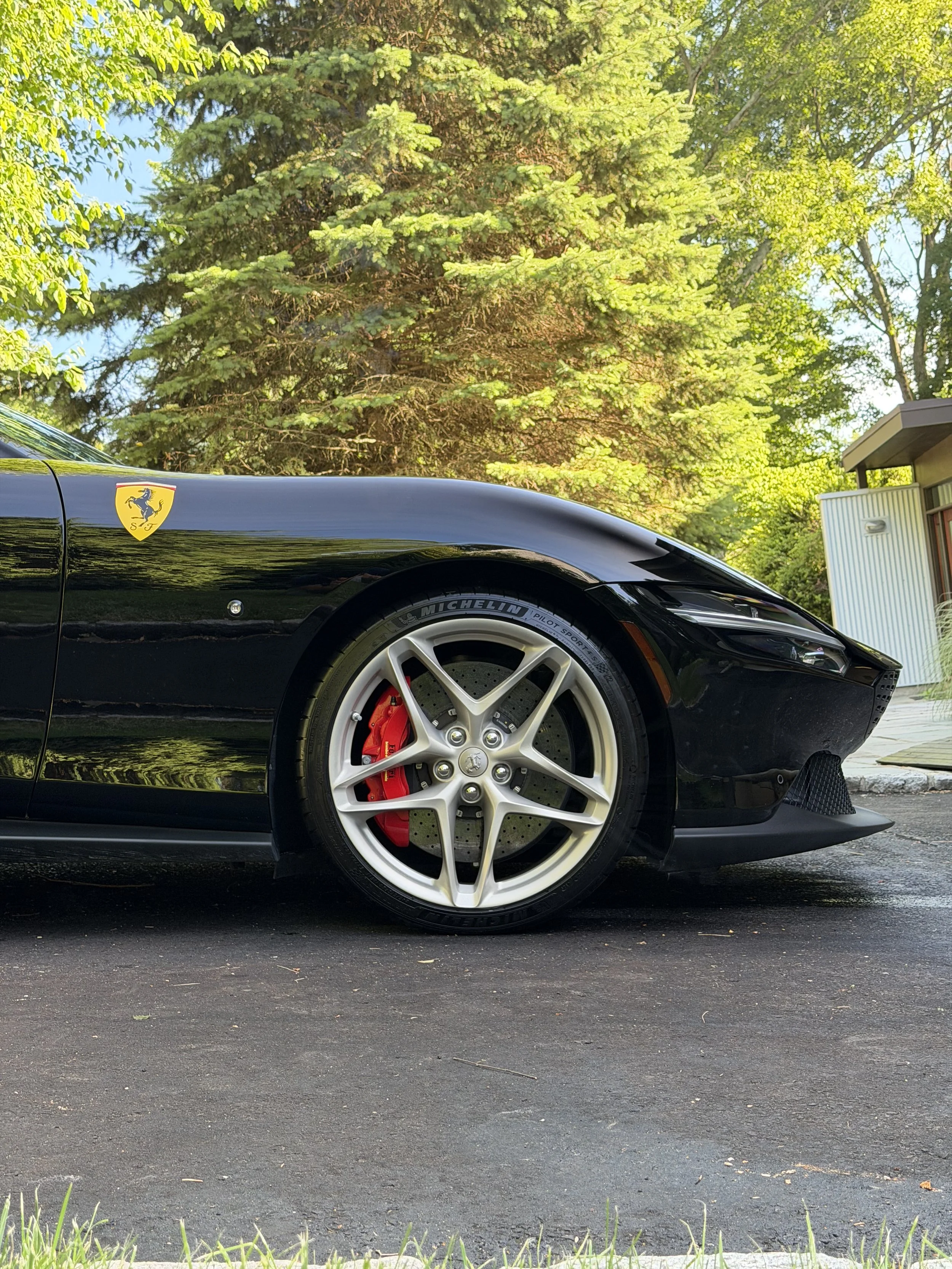 A black sports car with a Ferrari emblem on the side, parked on a paved road with green trees and a building in the background.