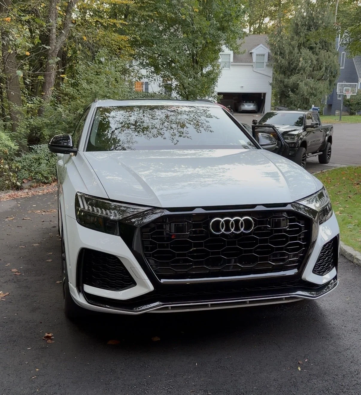 A white Audi sedan parked on a driveway surrounded by trees and houses, with two black trucks visible in the background.