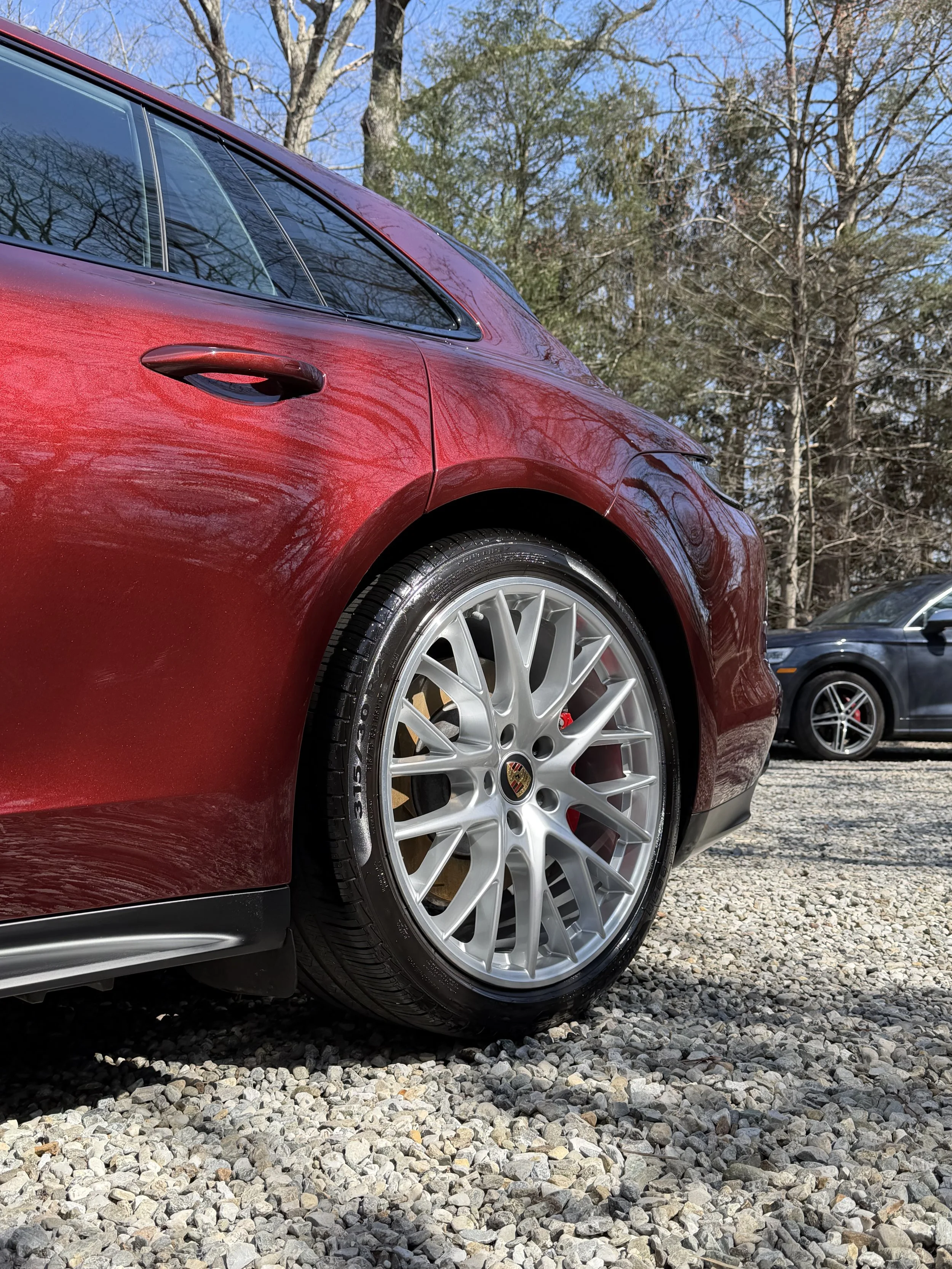 Close-up of a red sports car's front side, including wheel, body, and part of the windshield, with trees and blue sky in the background.