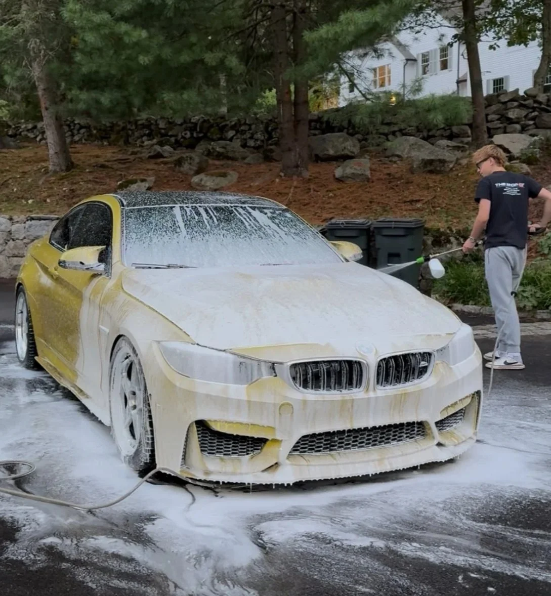 A young man is washing a yellow sports car with soap suds and water outside during daytime. The car is covered in soap and is being rinsed off with a hose. There are trees, rocks, and a white house in the background.