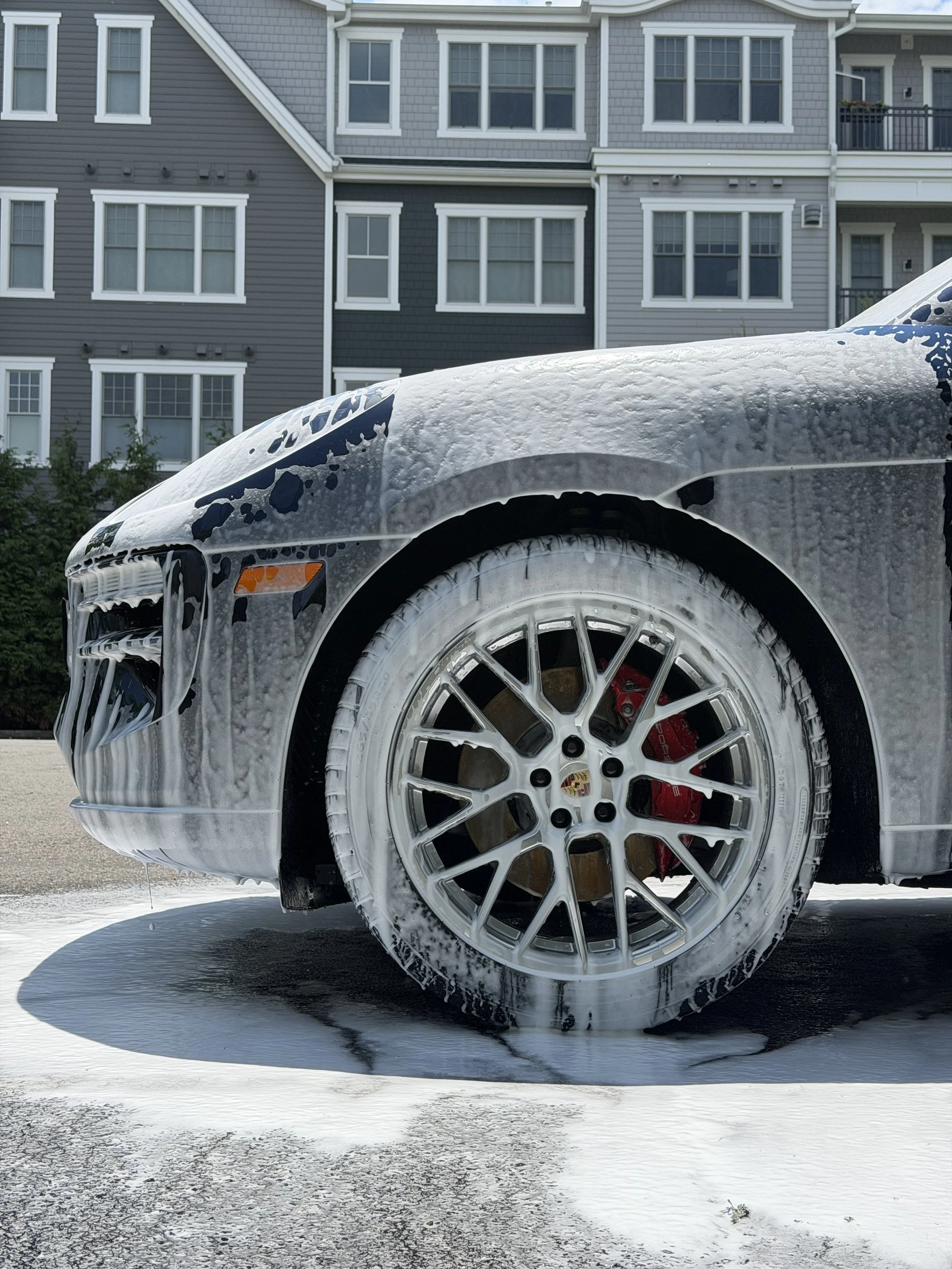 Close-up of a black luxury car with soap suds on its front fender, wheel, and tire, parked outdoors with a shadow and a multi-story residential building in the background.