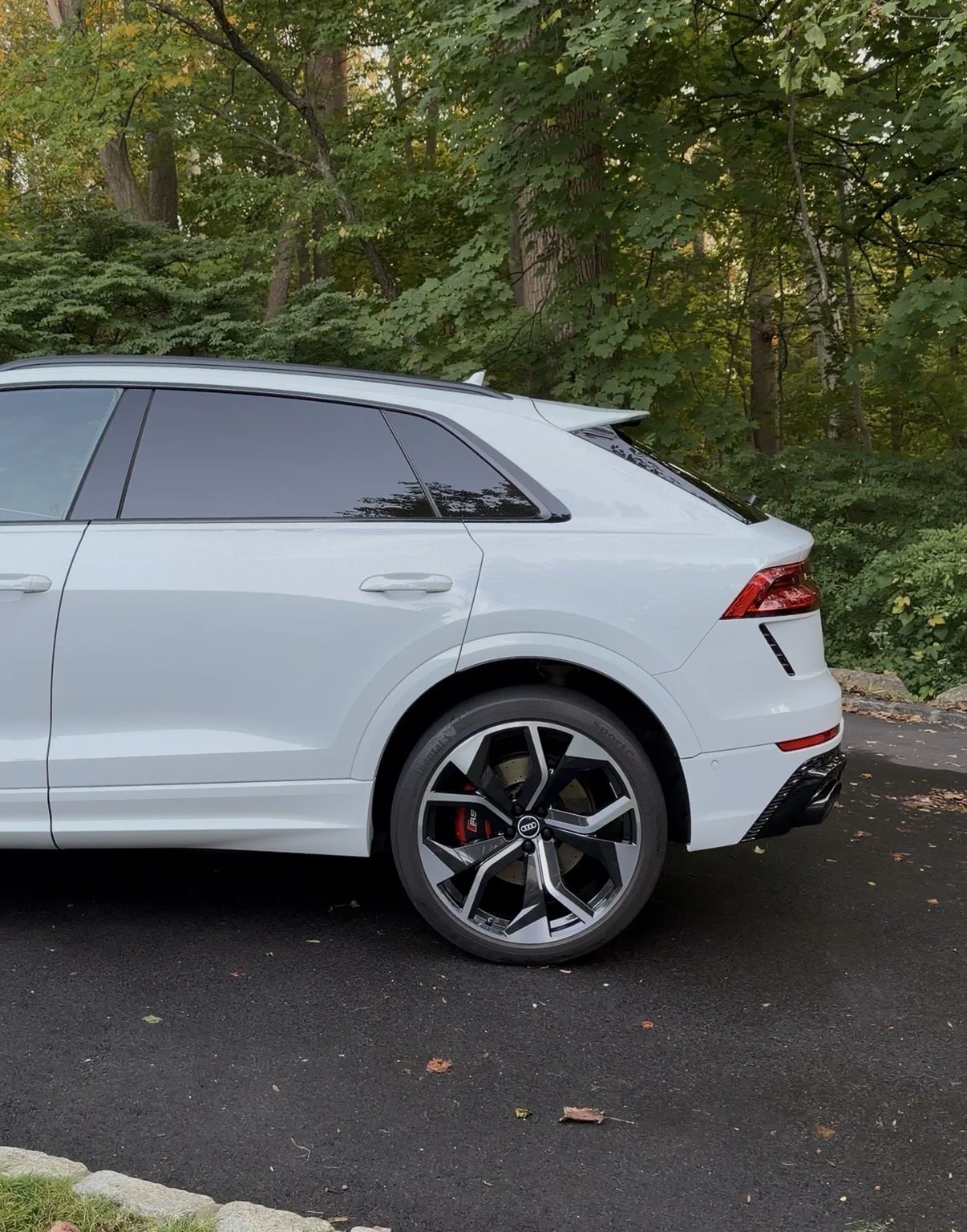 The rear half of a white Audi RS SUV with black alloy wheels and tinted windows parked on a paved surface in front of a wooded area.