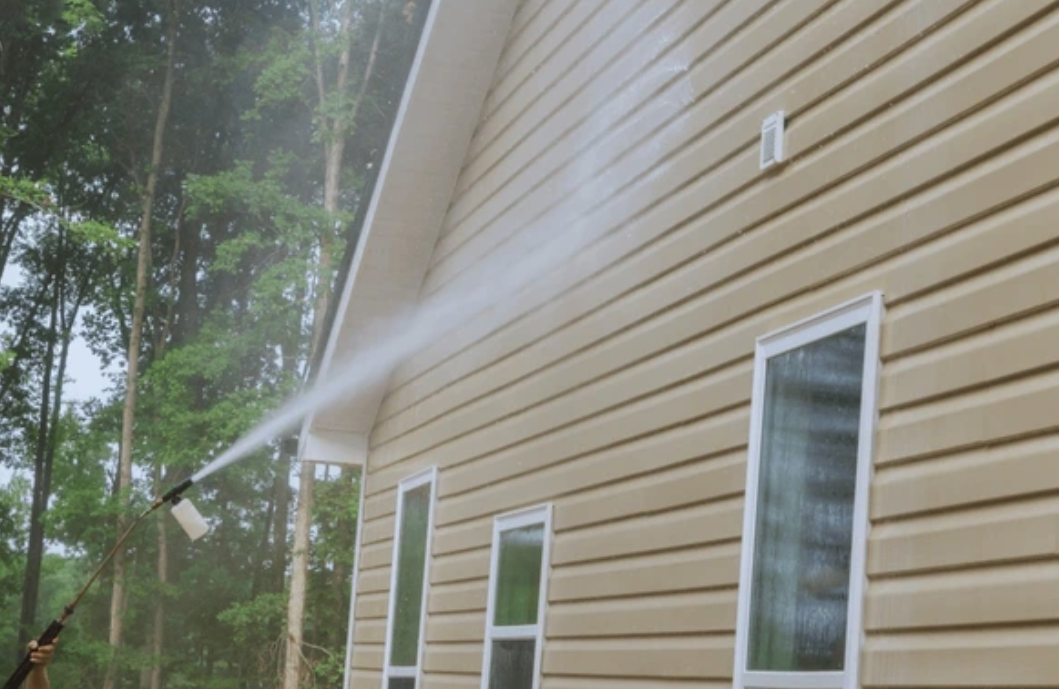 A person power washing the beige siding of a house with water spraying from the nozzle, near tall trees.