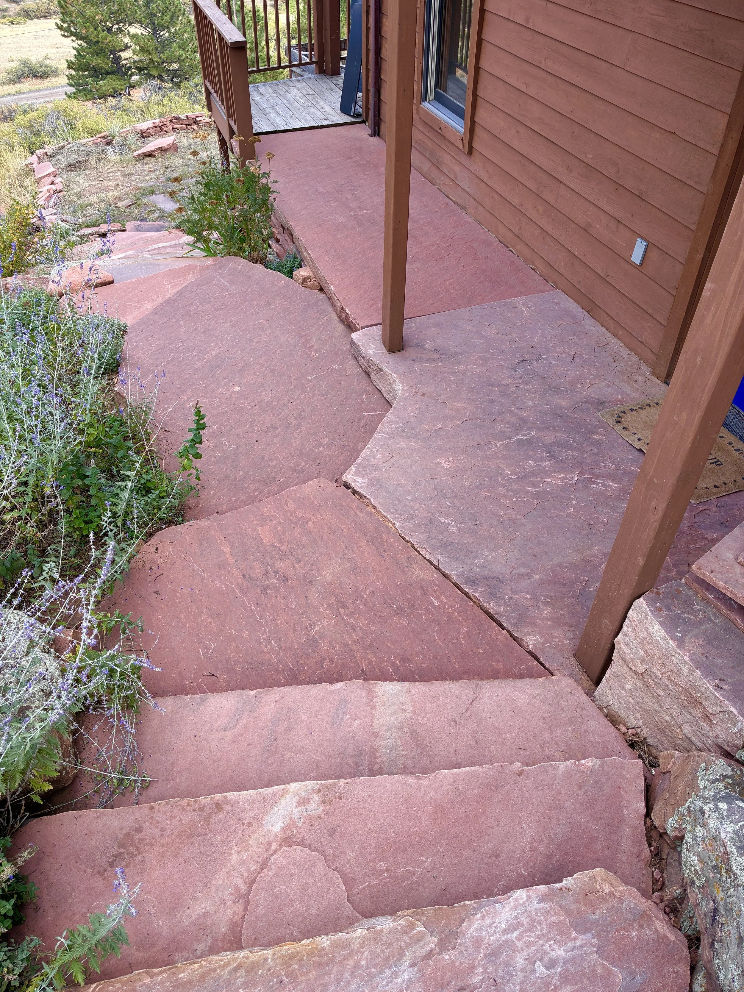 Red sandstone steps leading up to a wooden deck porch with a brown wood exterior house wall and a small plant at the side, surrounded by some greenery and landscape in the background.