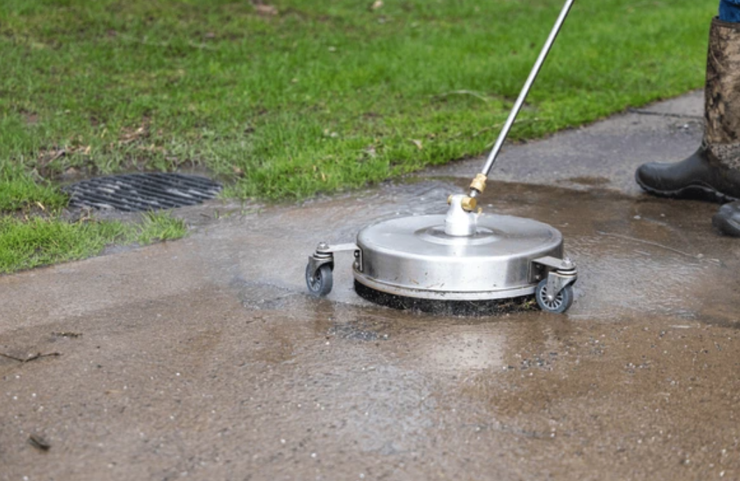A person power washing a concrete sidewalk near grass, wearing camo-patterned boots.