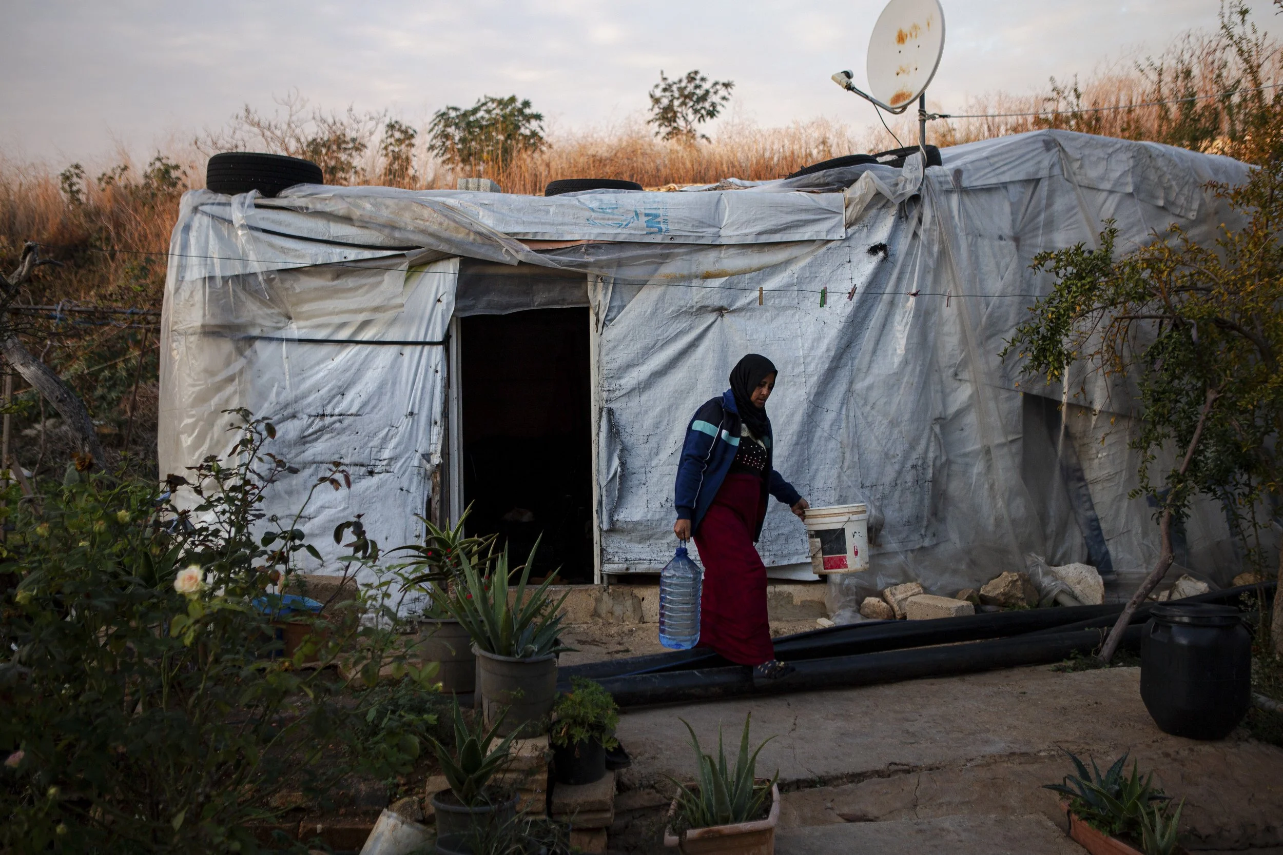 Ghazieye lifts a container of water from her tented shelter to bring to the kitchen, where her sister is cooking. She and her sisters built the shelter more than 10 years ago using UNHCR canvas, plastic sheets, tires, and wood — a space originally ma