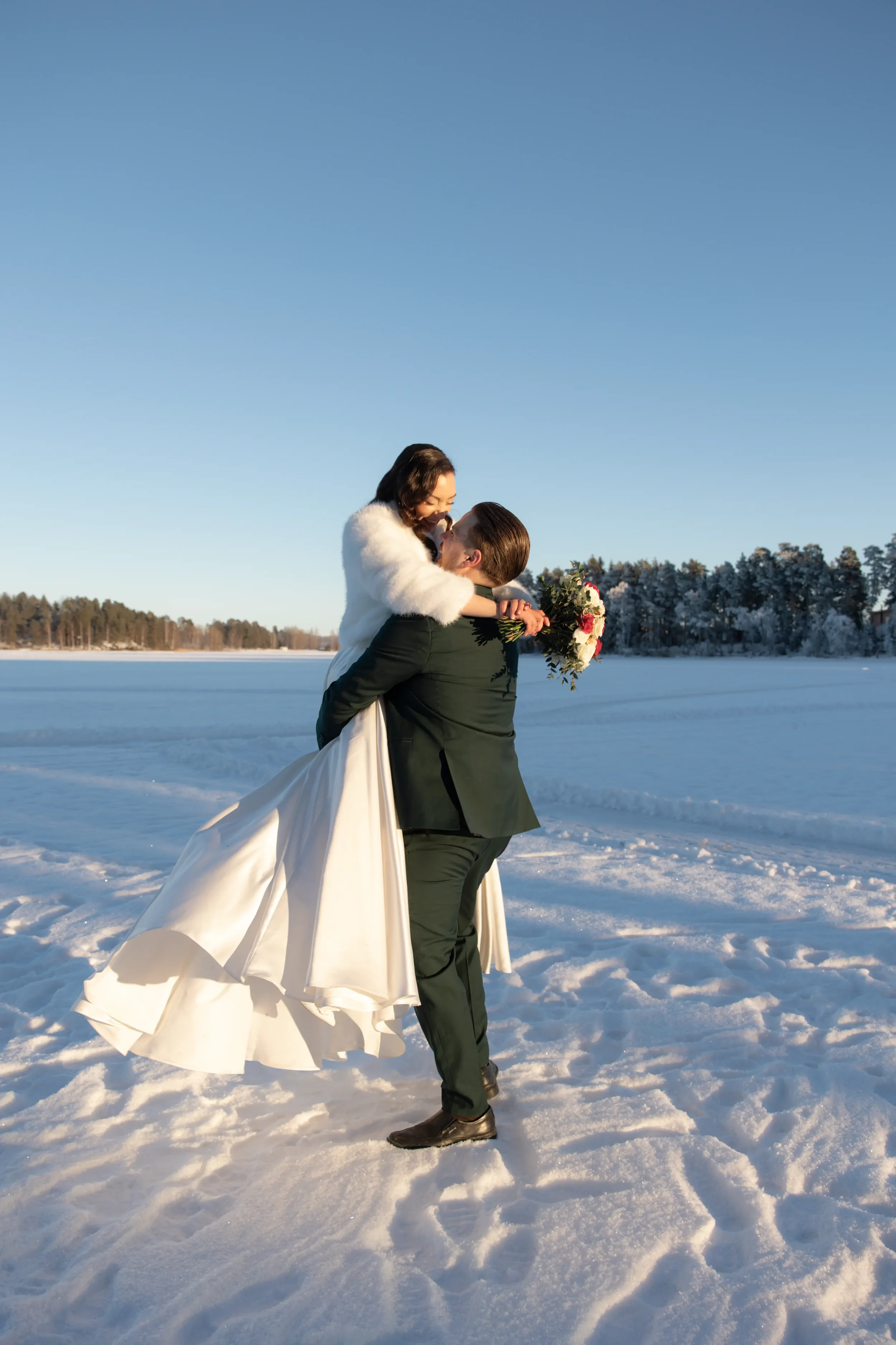 Hääpari lumisella jäällä, mies nostaa morsiamen ilmaan ja he katsovat toisiaan rakastuneina, taustalla talvinen maisema ja sininen taivas. Hääkuvaaja Tampere.