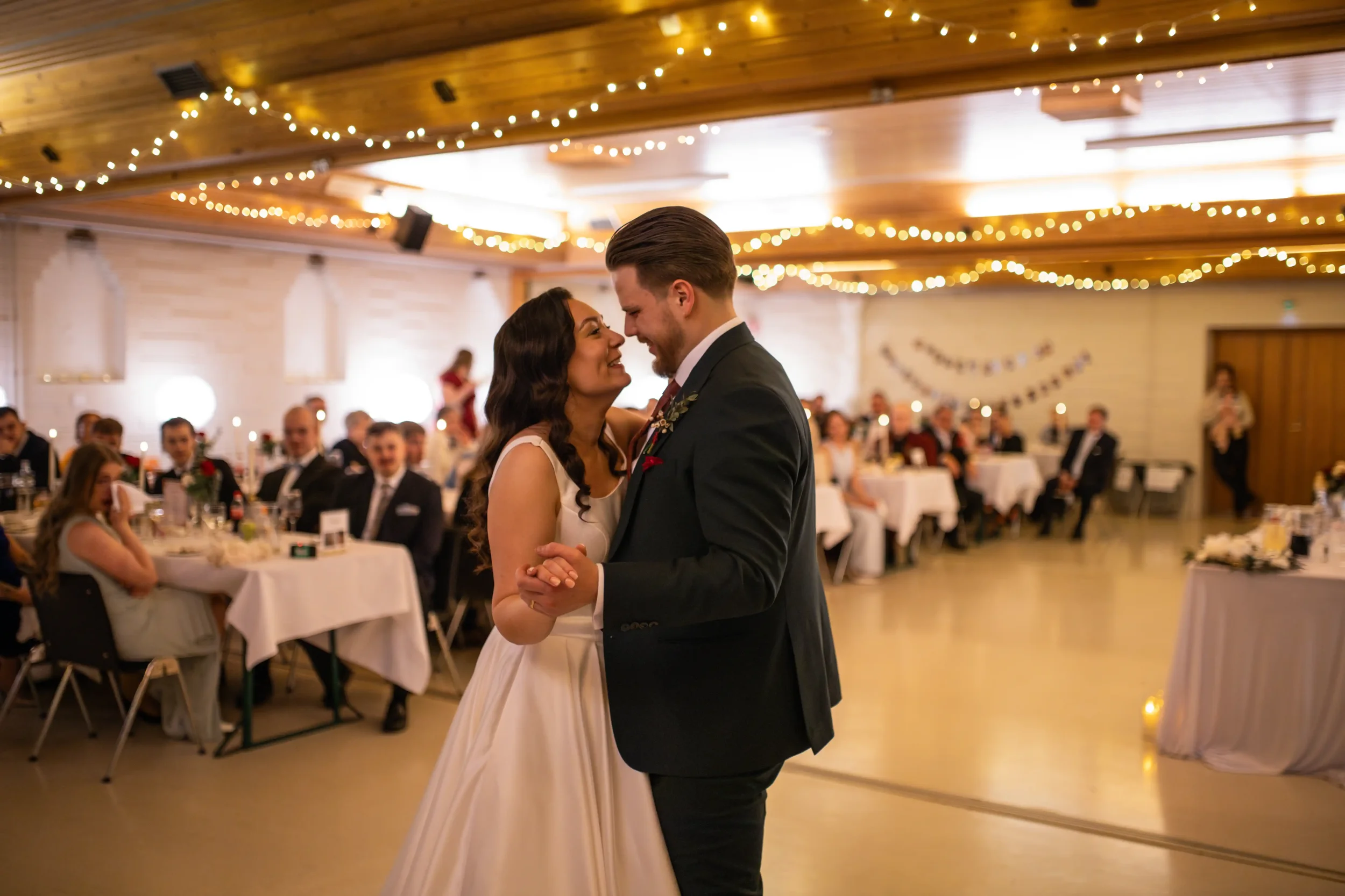 Hääpari tanssii keskellä juhlasalia, taustalla ihmisiä istumassa pöydissä ja ripustettuina valoja. Hääkuvaaja Tampere.