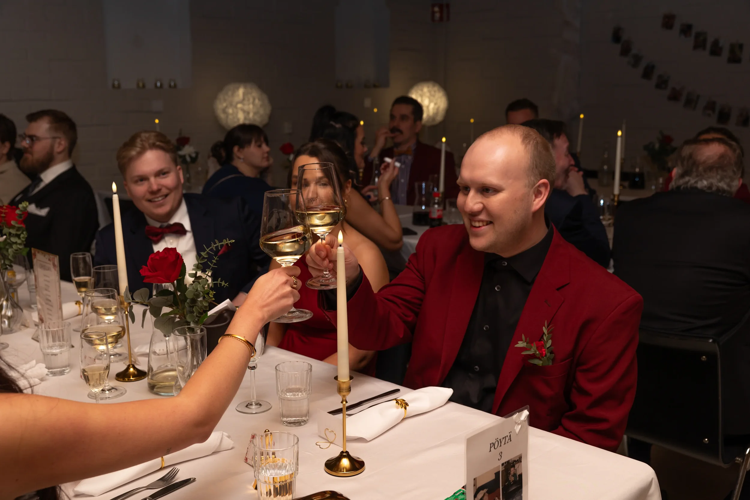 Ihmisiä juhlapöydässä kippistäen viinilaseilla häissä, jossa on kynttilöitä ja kukka-asetelmia. Hääkuvaaja Tampere.