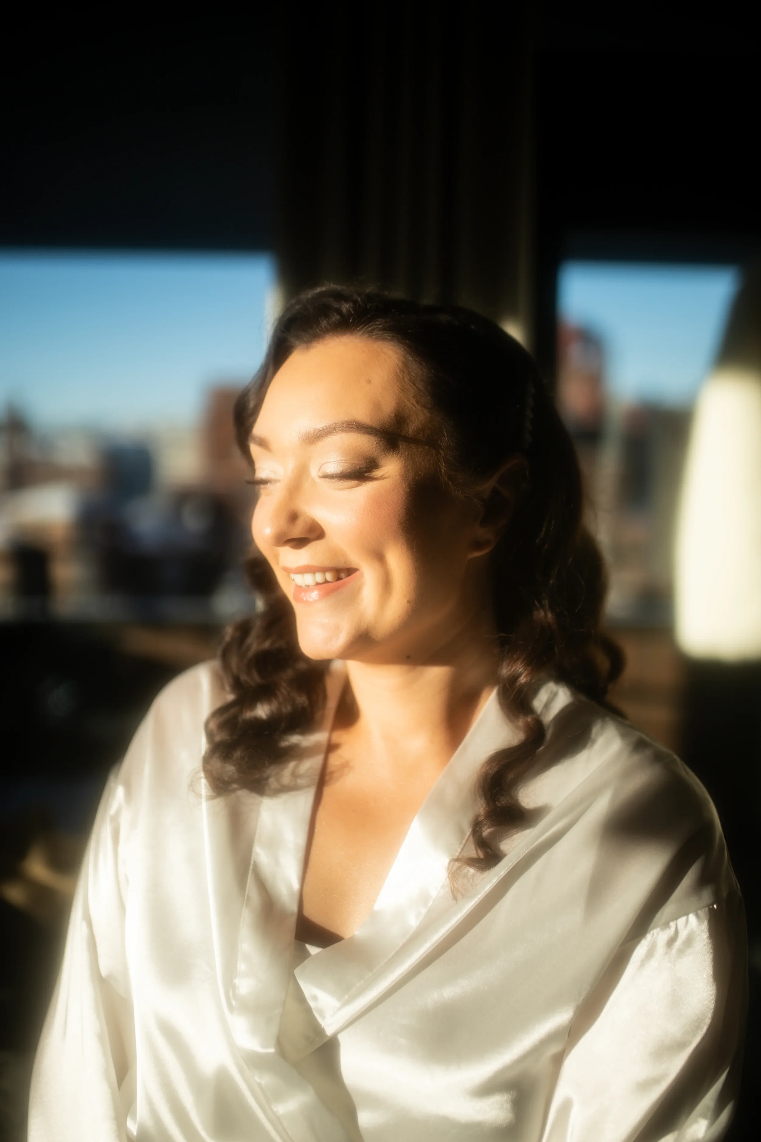 Morsian hymyilee ja istuu huoneessa auringonvalossa, valkoisessa satiiniaamutakissa. Hääkuvaaja Tampere