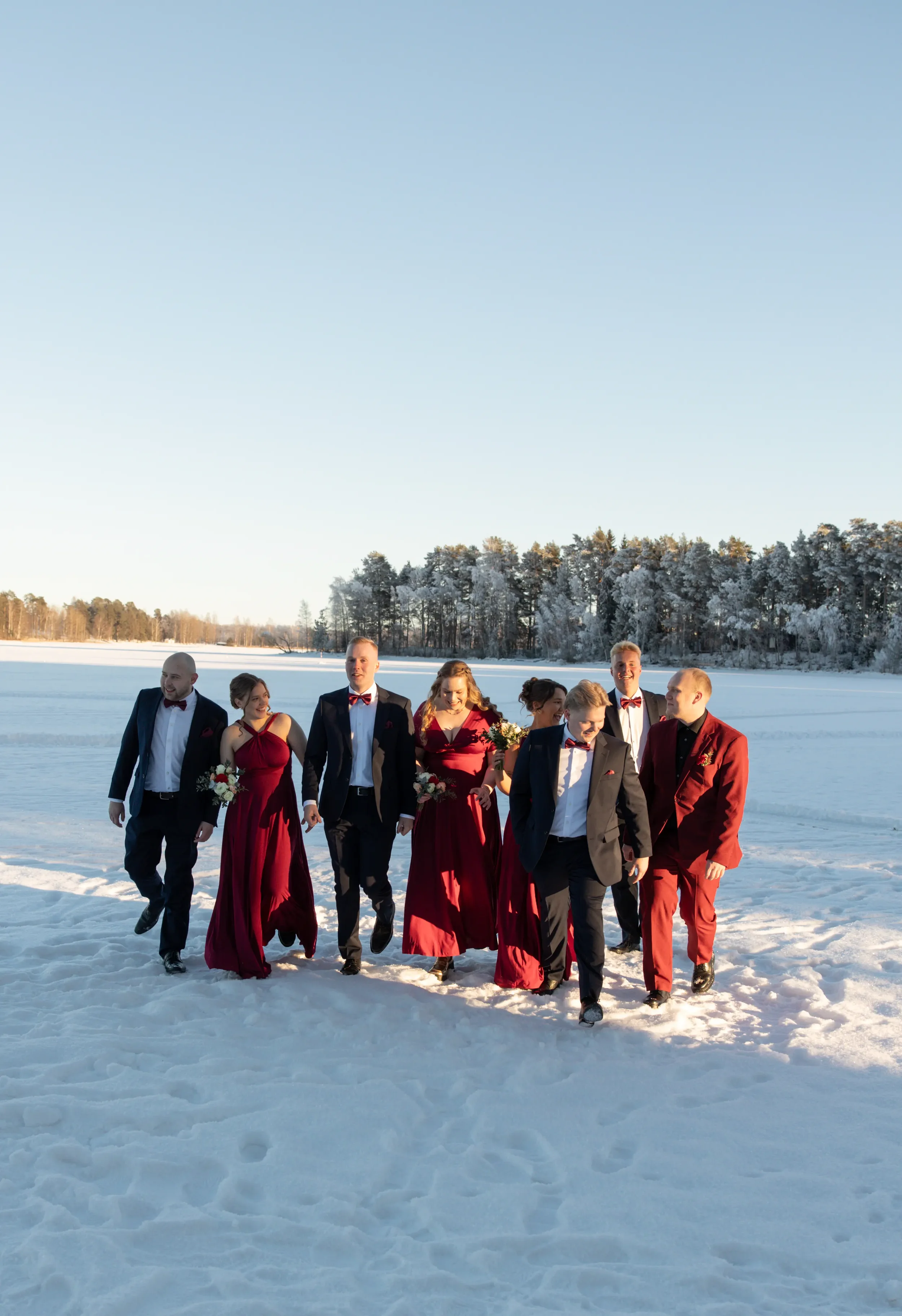 Hääpari sekä kaasot ja bestmanit kävelemässä lumisella järven jäällä talvella, taustalla jäiset puut ja selkeä taivas. Hääkuvaaja Tampere.