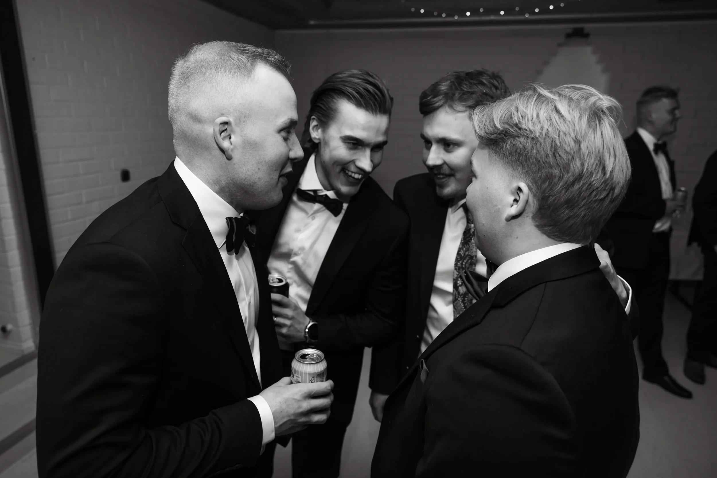 Mustavalkoinen kuva neljästä pukumiehestä juhlassa, keskustelemassa ja nauttimassa alkoholia. Hääkuvaus Tampere.