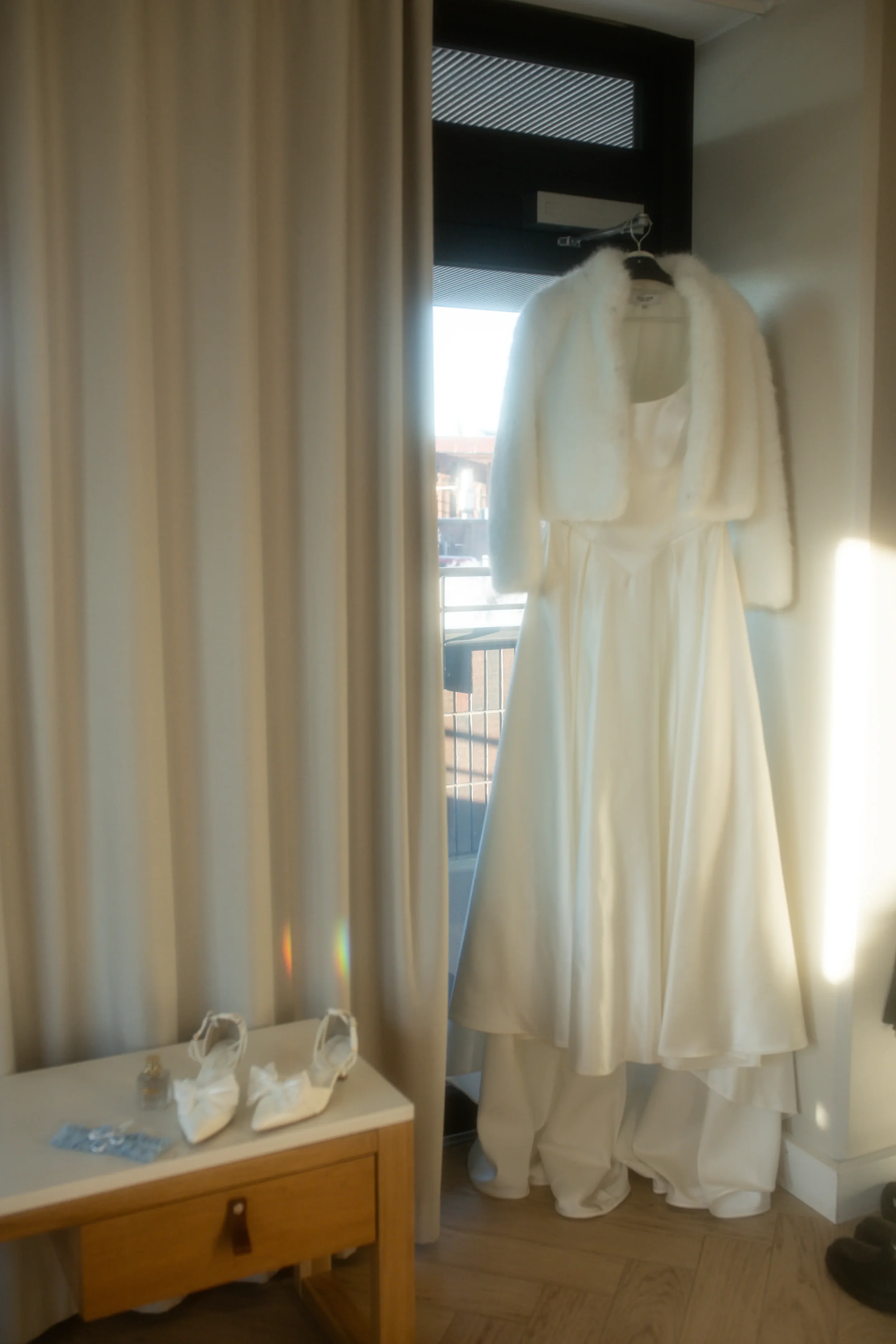 Häämekko ja karvatakki roikkumassa ikkunan edessä, jalkineet ja hajuvesi pöydällä vieressä. Hääkuvaaja Tampere.