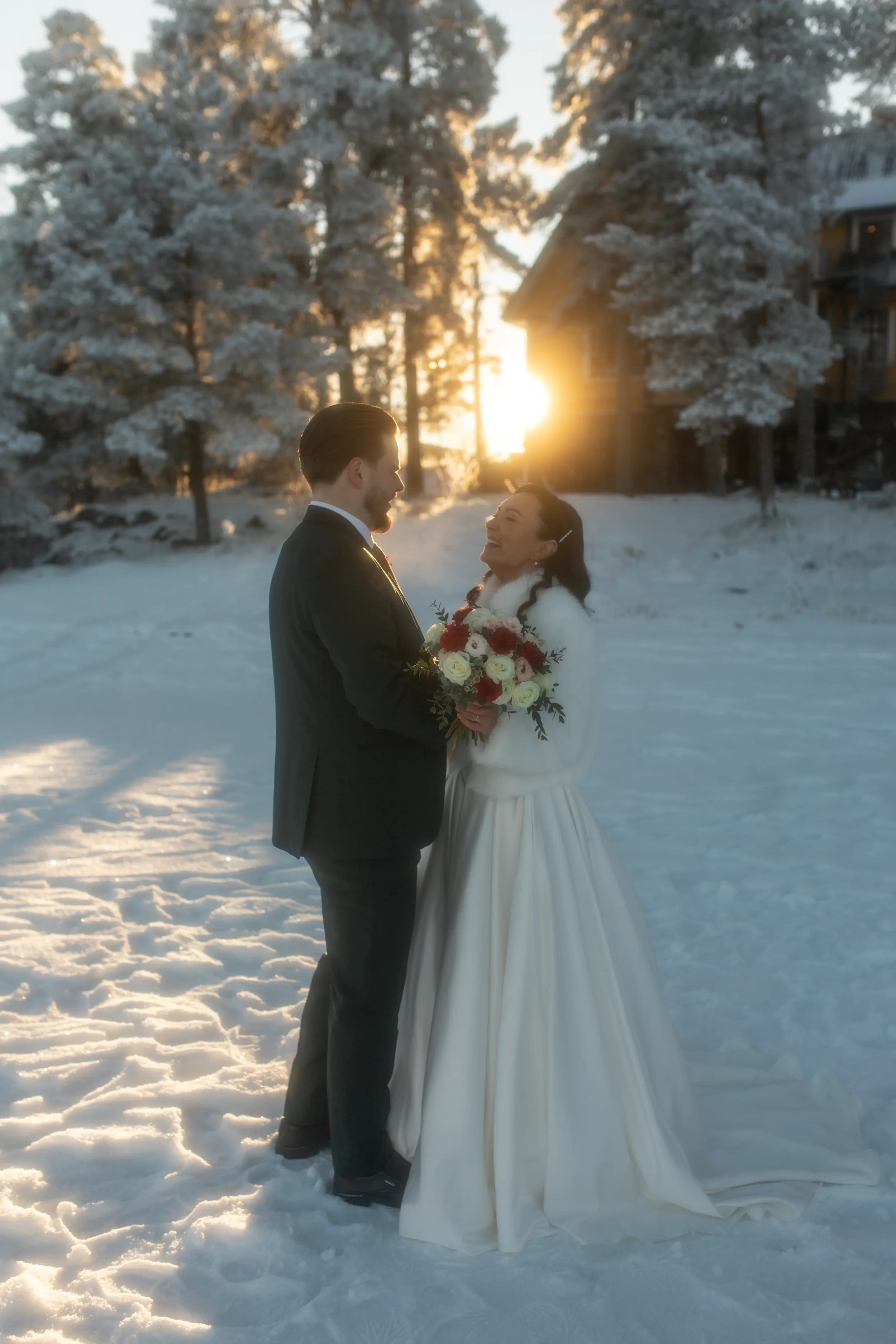 Hääpari juttelee lumisessa maisemassa auringonlaskun aikaan, nainen pitää kukkakimppua, taustalla lumiset puut ja rakennus. Hääkuvaaja Tampere.
