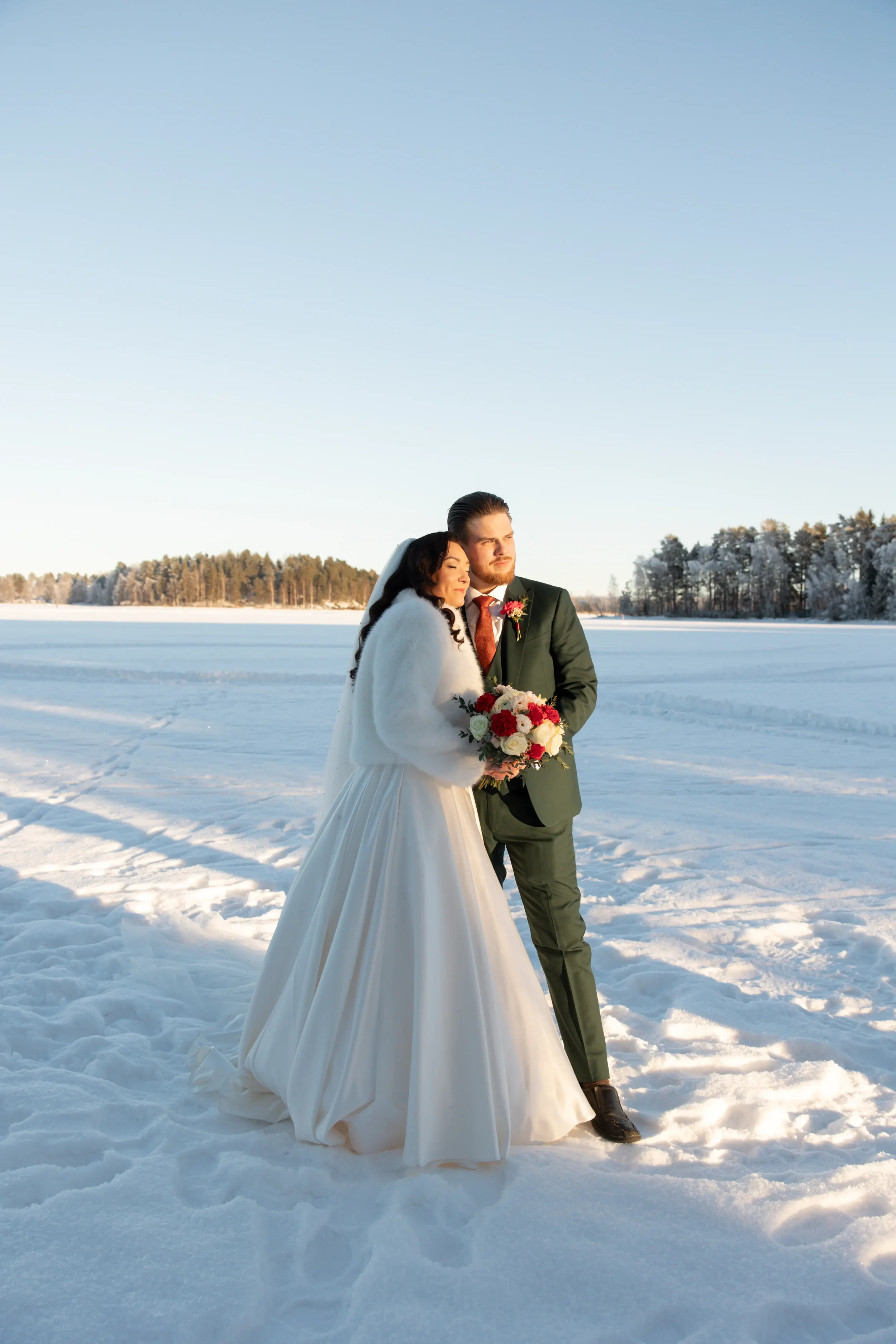 Hääpari seisoo lumisessa maisemassa, nainen on valkoisessa hääpuvussa ja mies pikkutakin ja solmion kanssa, nainen pitää kukkakimppua. Taivas on kirkas ja taustalla on järvi ja lumiset metsät. Hääkuvaaja Tampere.
