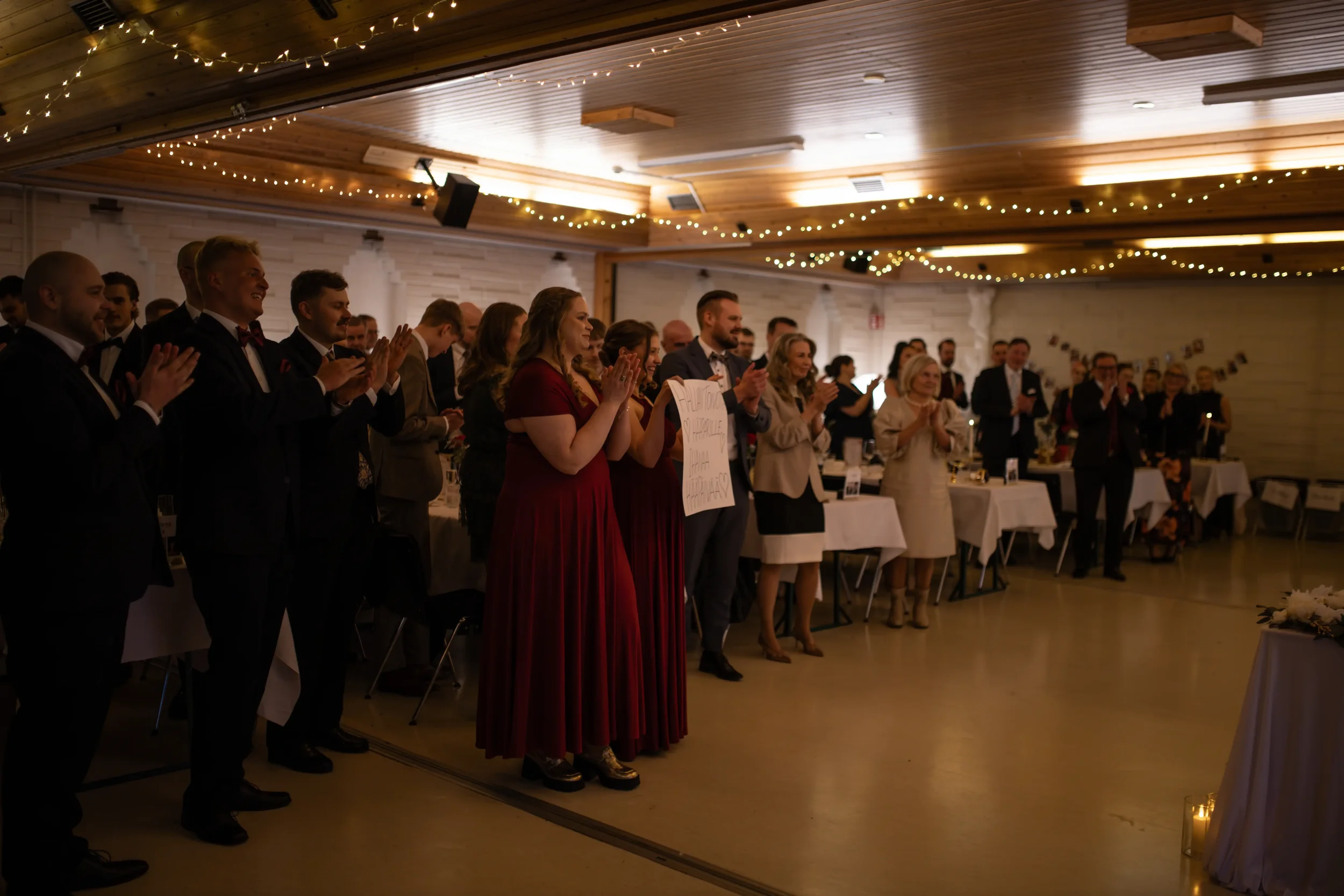 Juhlaväkeä seisomassa sisätiloissa, hymyilevät ja taputtavat, juhlapuvuissaan, pöytiä ja kynttilöitä, koriste- ja valoketjuja katossa. Hääkuvaaja Tampere.
