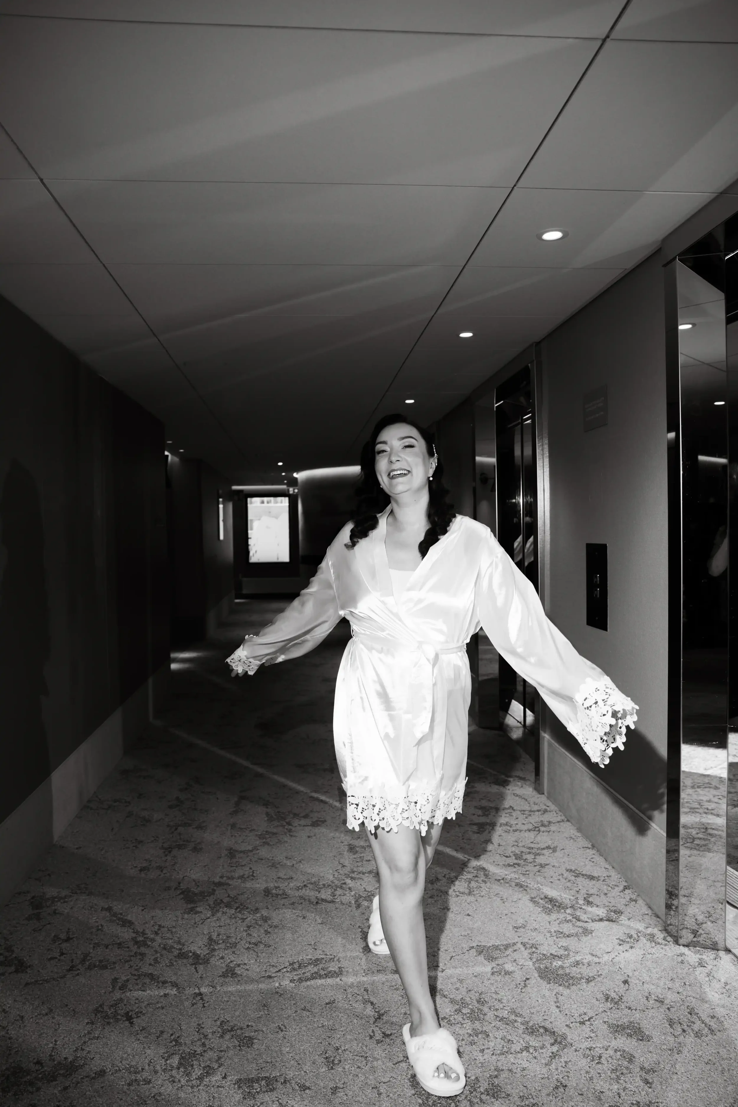 Nainen kävelee hotellin käytävällä, nauttii, hymyilee ja on pukeutunut valkoiseen satiiniaamutakkiin. Hääkuvaaja Tampere.