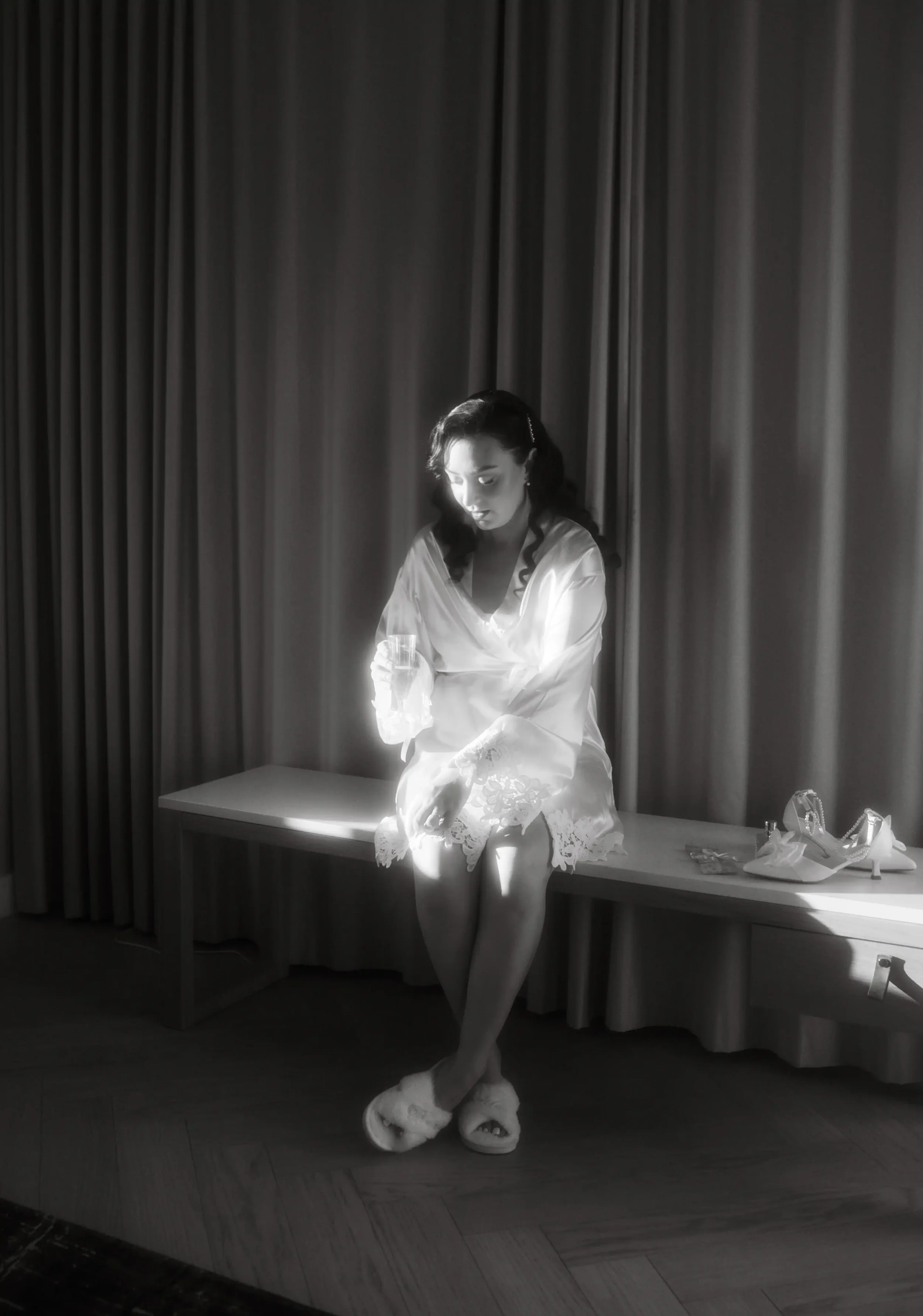 Morsian istuu verhon edessä, hohtaa valosta, pitäen lasia juomaa, yllään aamutakki ja korkokengät vieressään. Hääkuvaaja Tampere.