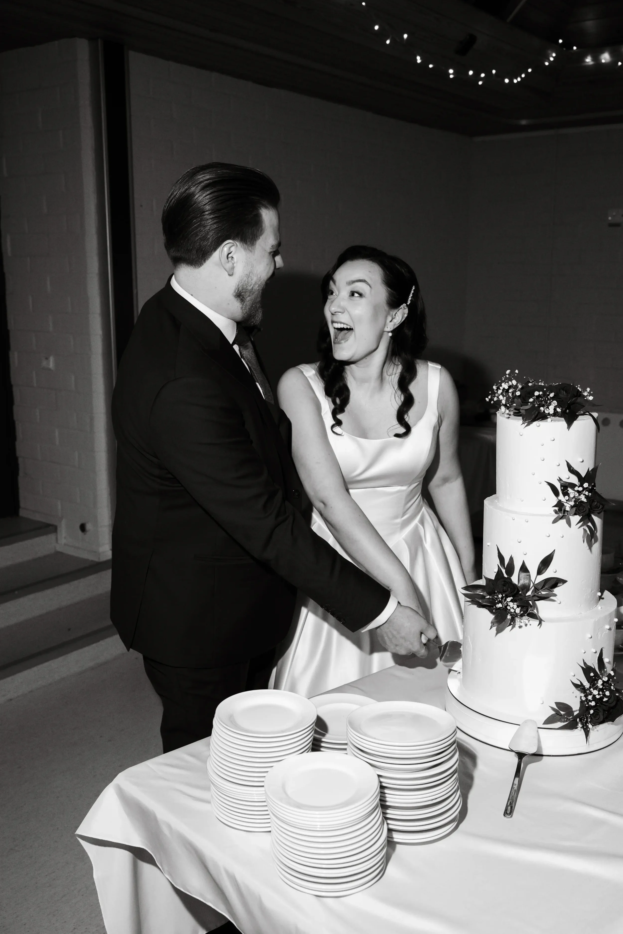 Hääpari leikkaa hääkakkua, jonka vieressä on paljon lautasia. Hääkuvaaja Tampere.