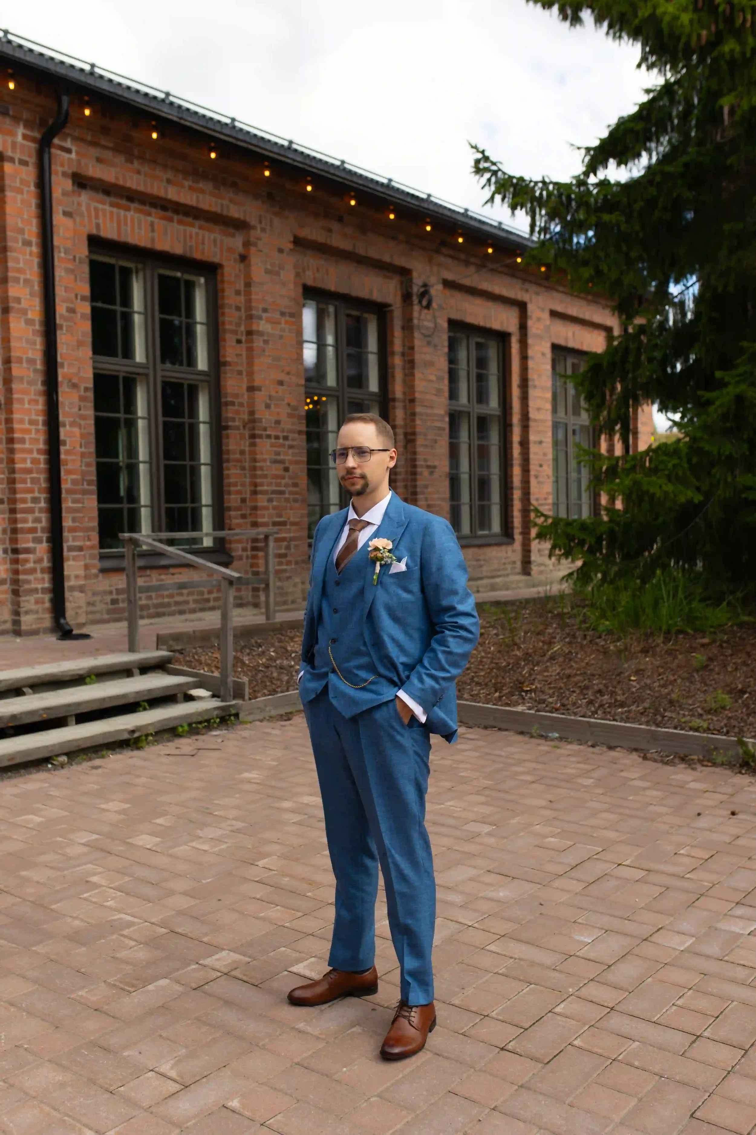 Mies hääpuvussa seisoo ulkona matalalla tiiliseinäisen rakennuksen edessä, jonka ikkunoista heijastuu sisävalaistus. Hänellä on sininen pikkutakki, raidallinen kravatti ja ruskeat kengät, ja hänellä on rintataskussa. Taustalla on vihreä havupuu ja ki