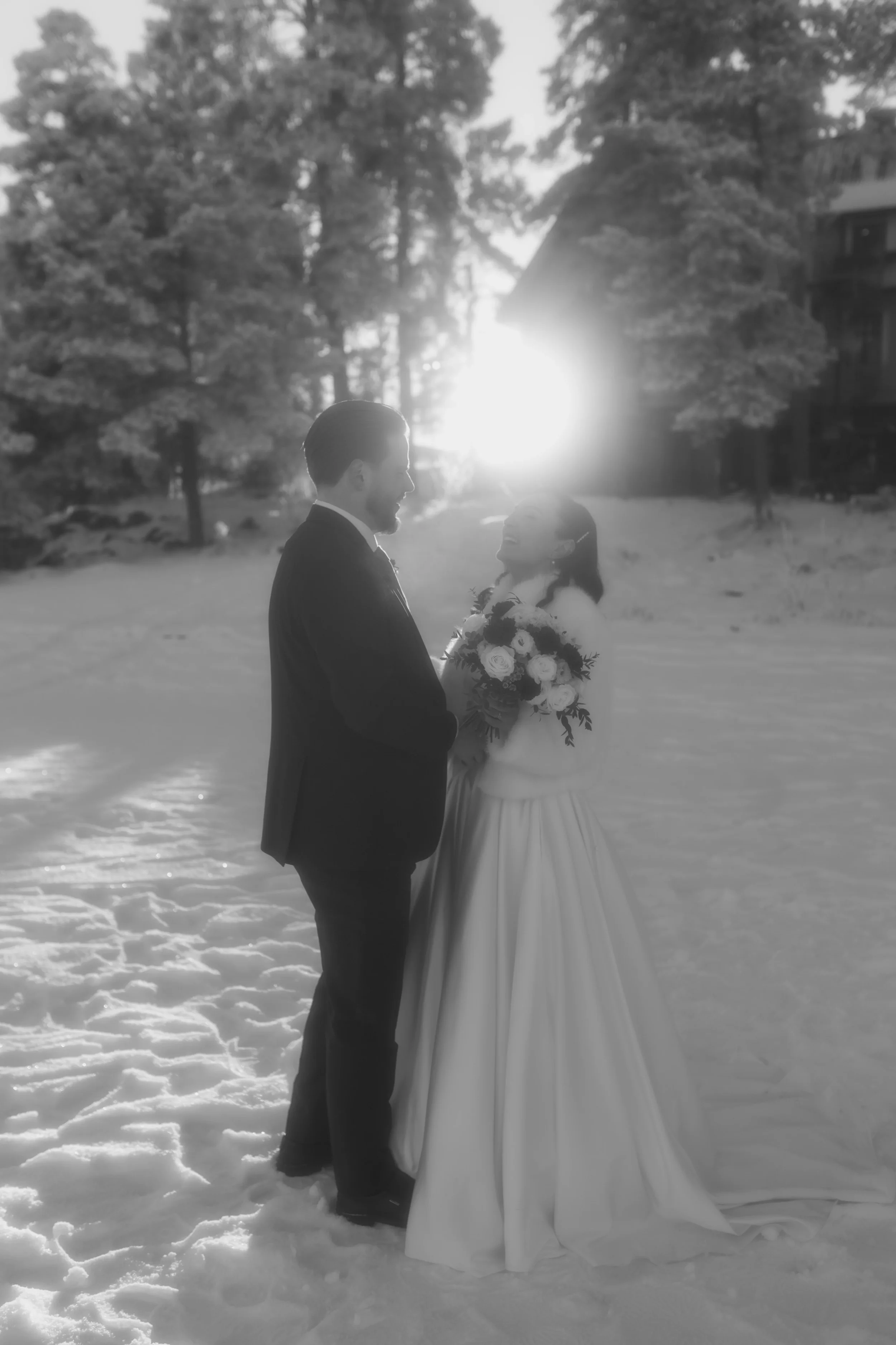 Hääpari seisoo lumisella alueella metsän ja rakennusten taustalla, auringonlasku taustalla, mustavalkoinen kuva. Hääkuvaaja Tampere.