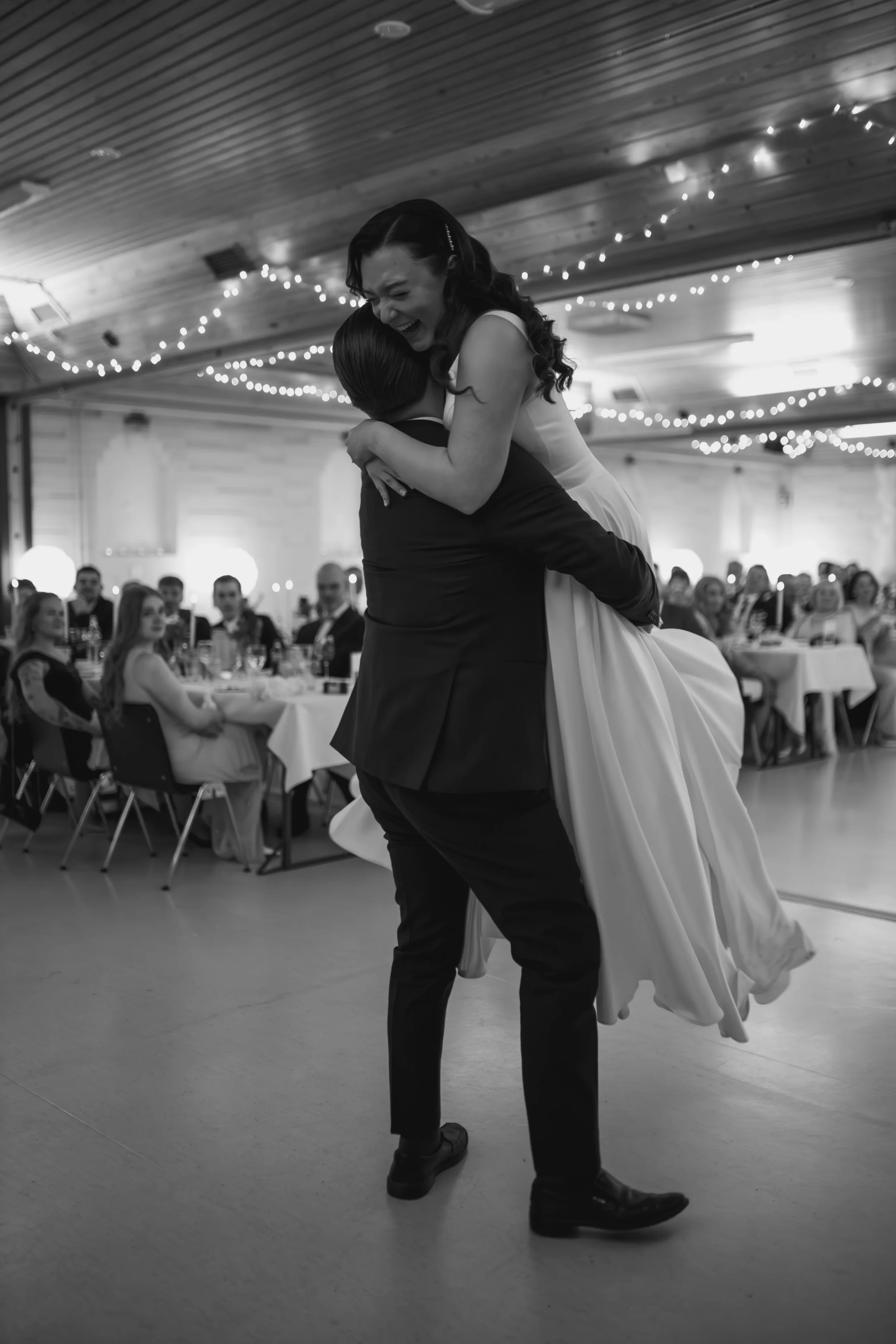 Morsiuspari tanssii hääparina, taustalla juhlayleisö istuu pöydissä. Tilassa on koristeellisia valoja ja puupanelikatto. Hääkuvaaja Tampere.