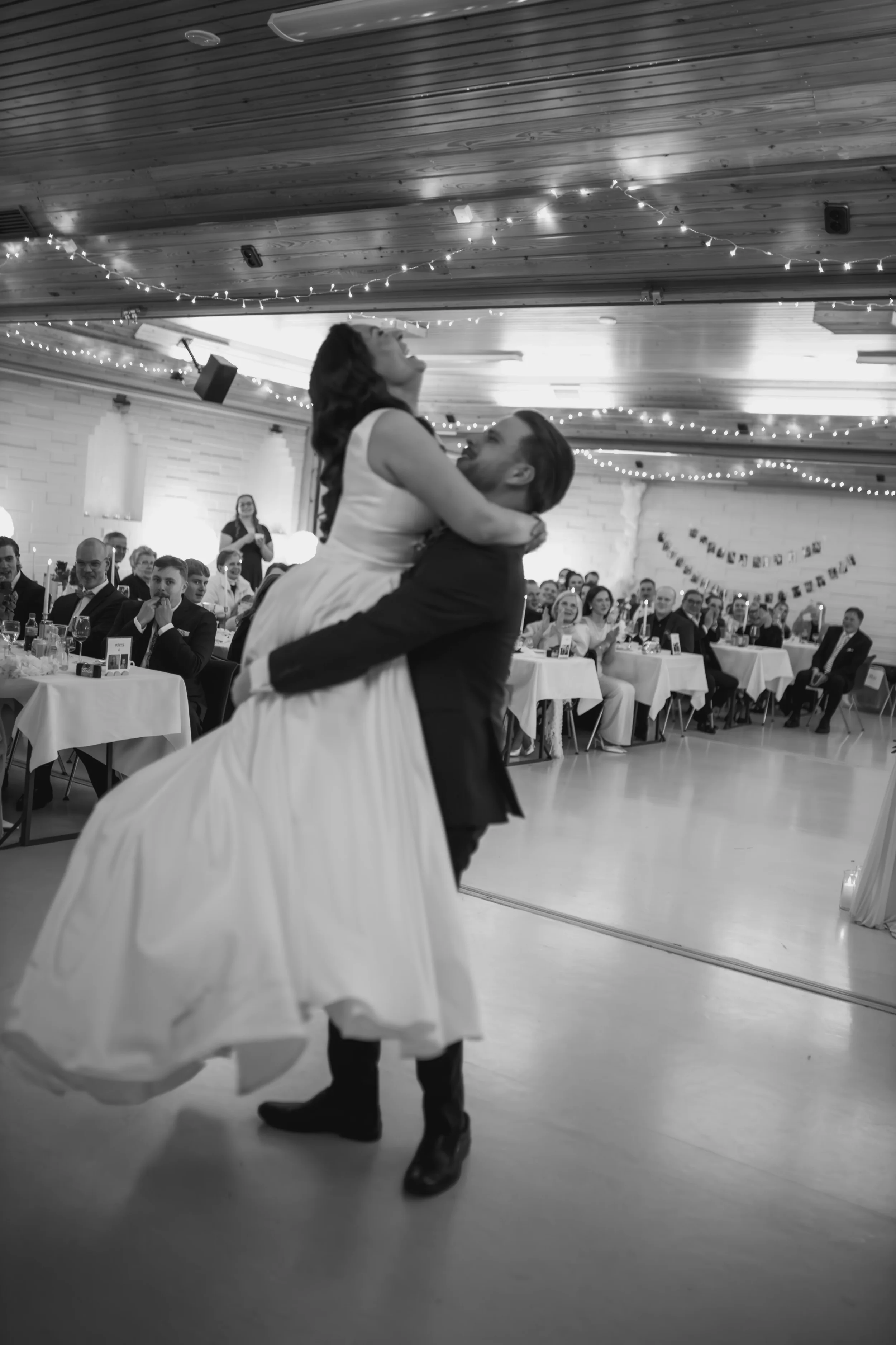 Häätanssi, jossa sulhanen nostaa morsiamen syliinsä, juhlapöydät ja vieraat taustalla. Hääkuvaaja Tampere.