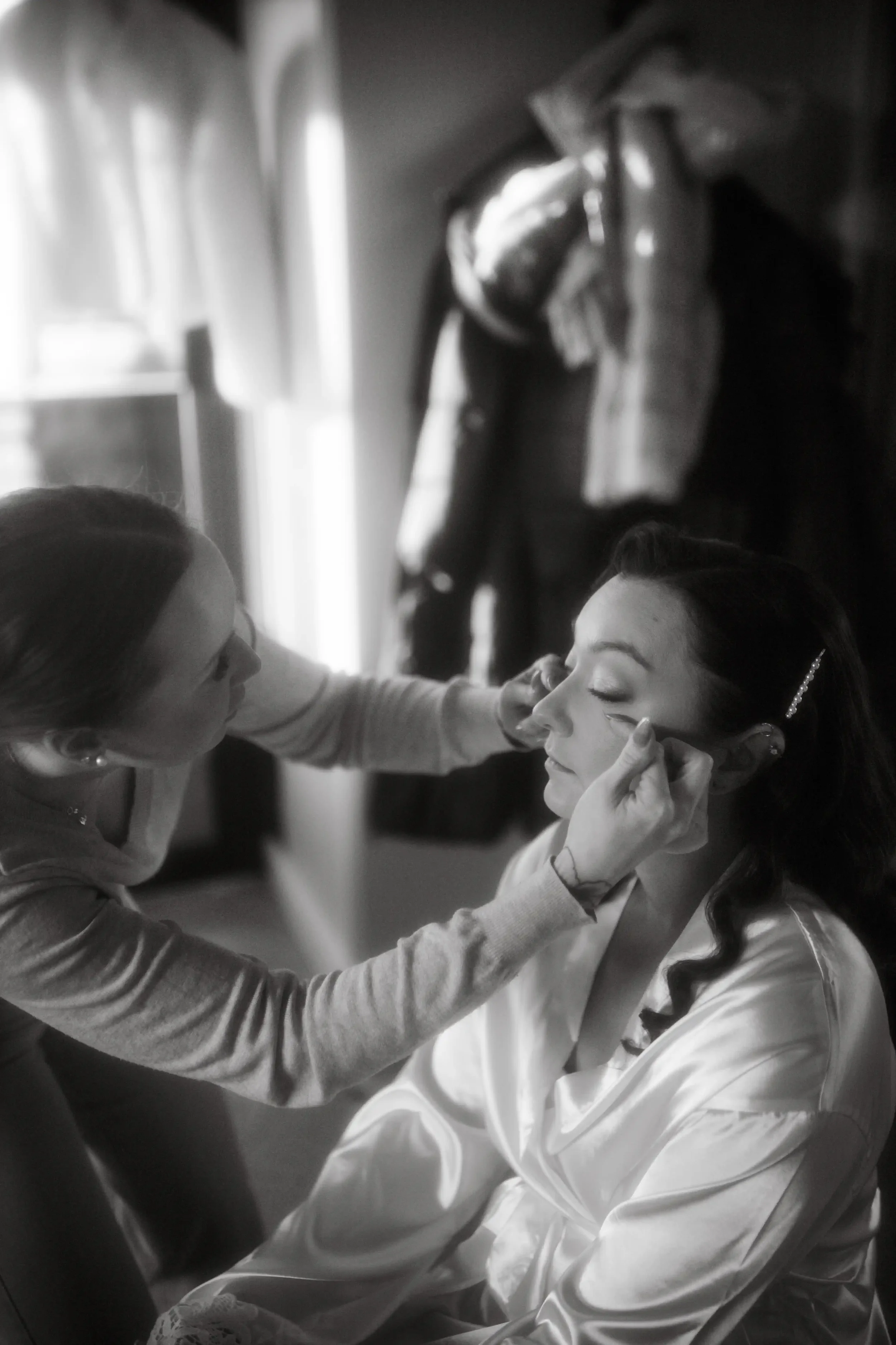 Nainen meikkaa morsianta. Morsiamella päällä valkoinen satiiniaamutakki. Taustalla vaatteita ja muita esineitä näkyvissä. Mustavalkoinen kuva. Hääkuvaaja Tampere.