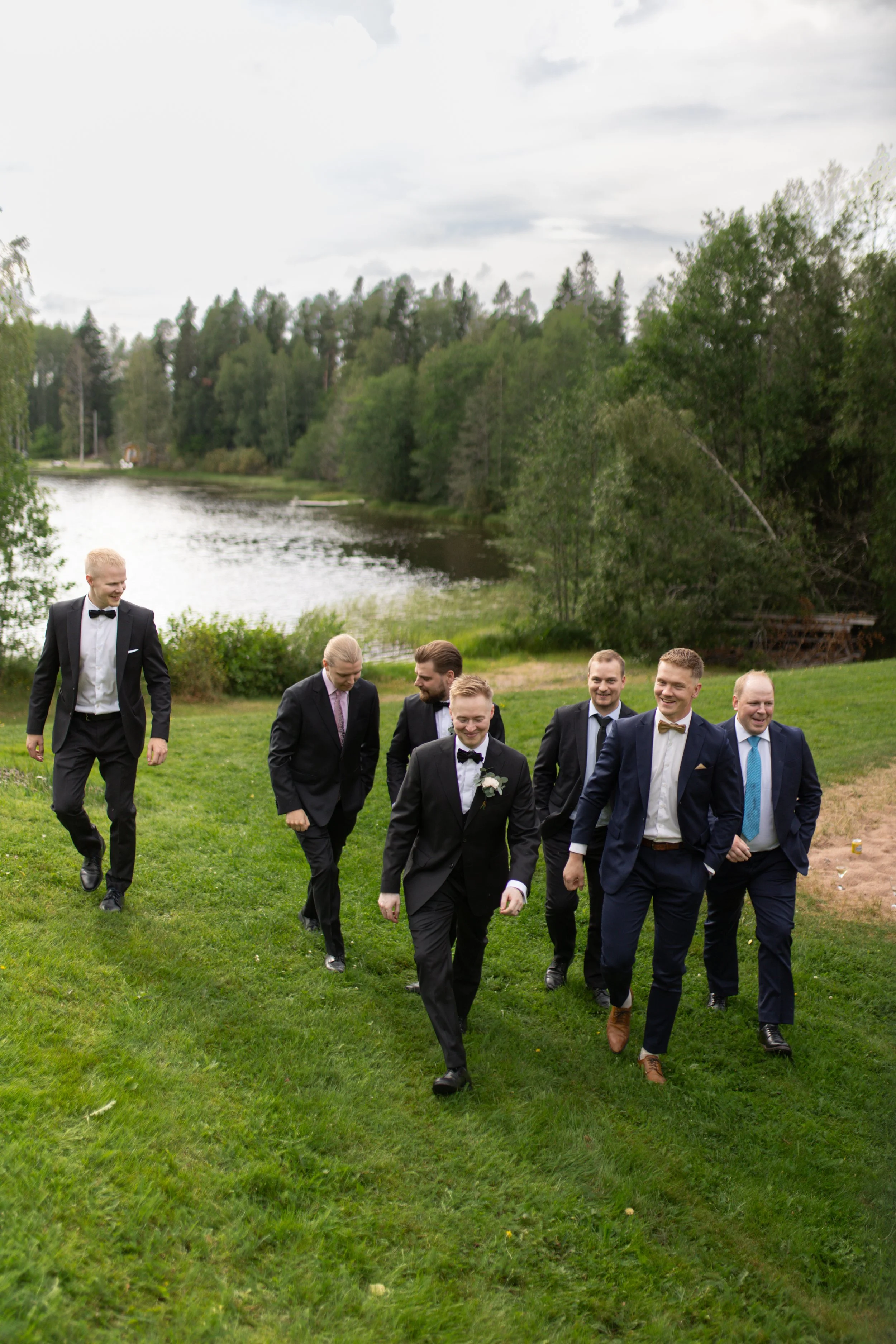 Mies porukka kävelee rinnettä ylös puvut päällä, vihreä nurmikko, järvi taustalla