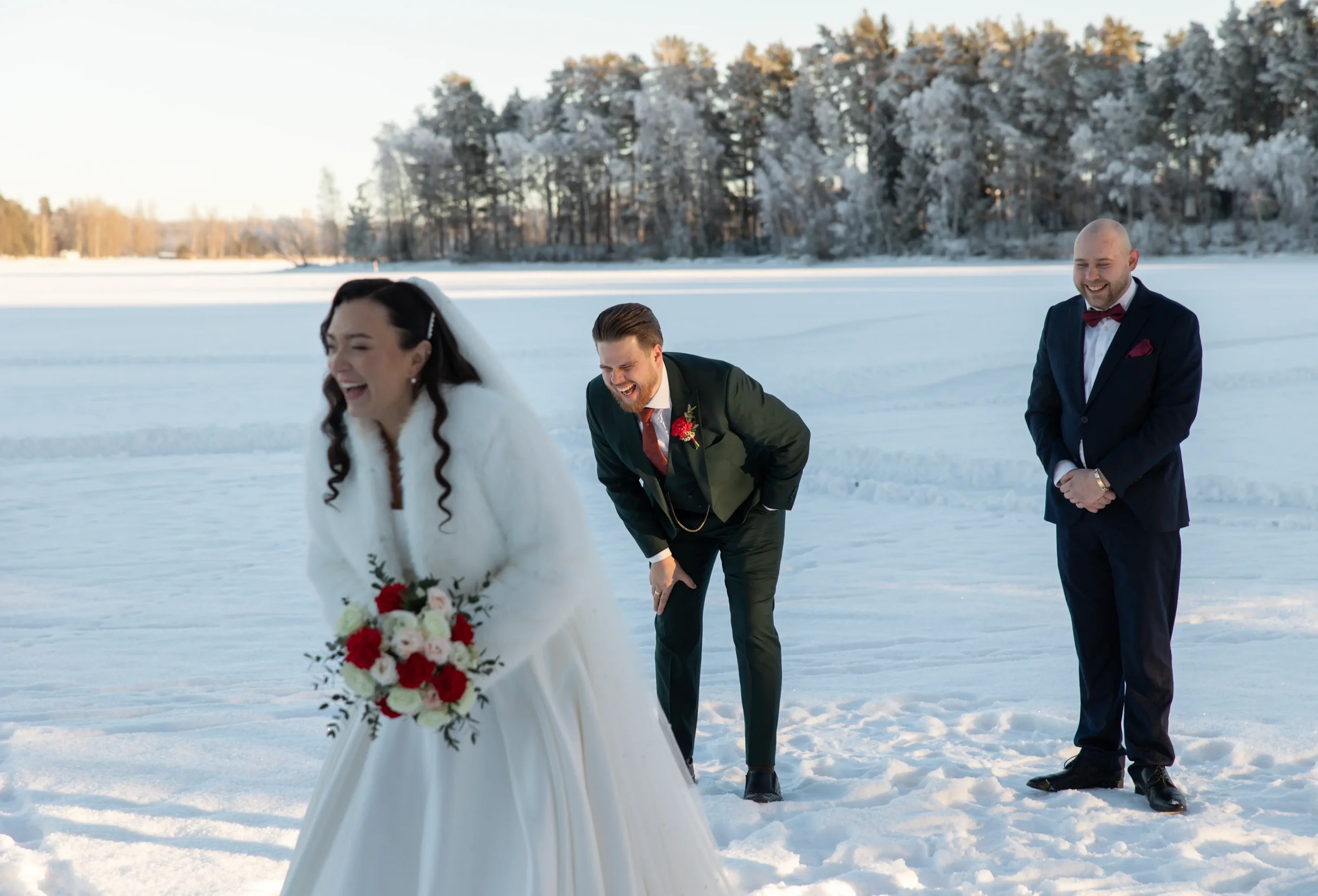 Hääpari ja ystävä nauravat lumisella järvenjäällä, taustalla luminen metsä ja sininen taivas. Hääkuvaaja Tampere.