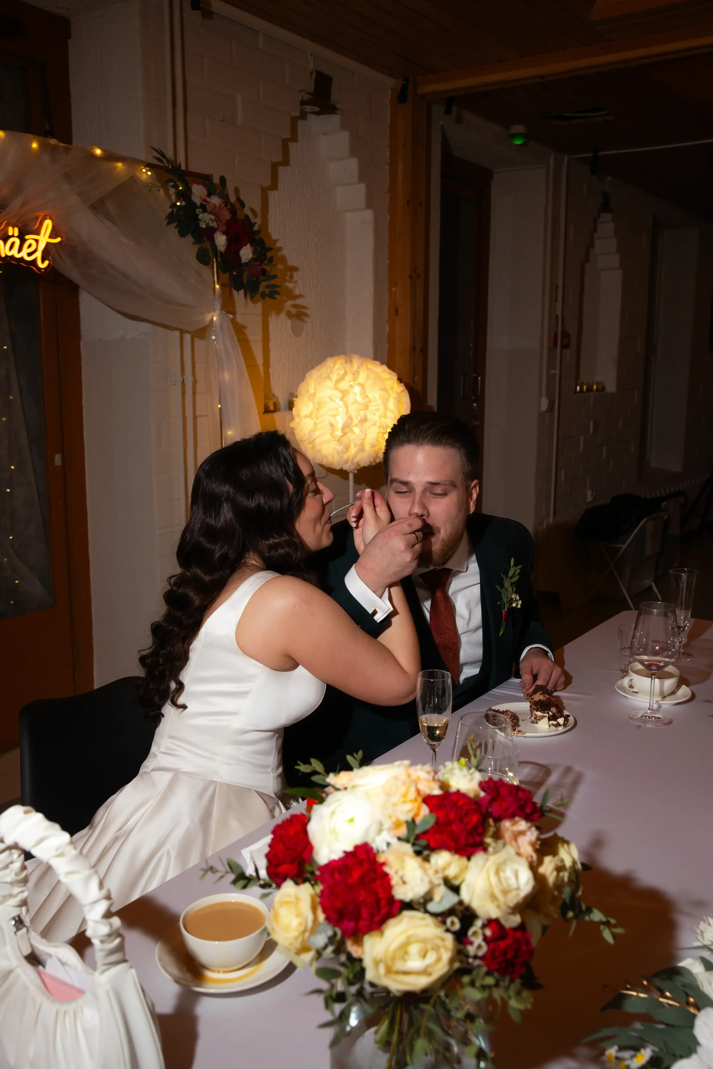 Hääpari nauttii kakusta ja juomista juhlapöydässä, ympärillä kukkakimppu ja juhlallisuudet. Hääkuvaaja Tampere.