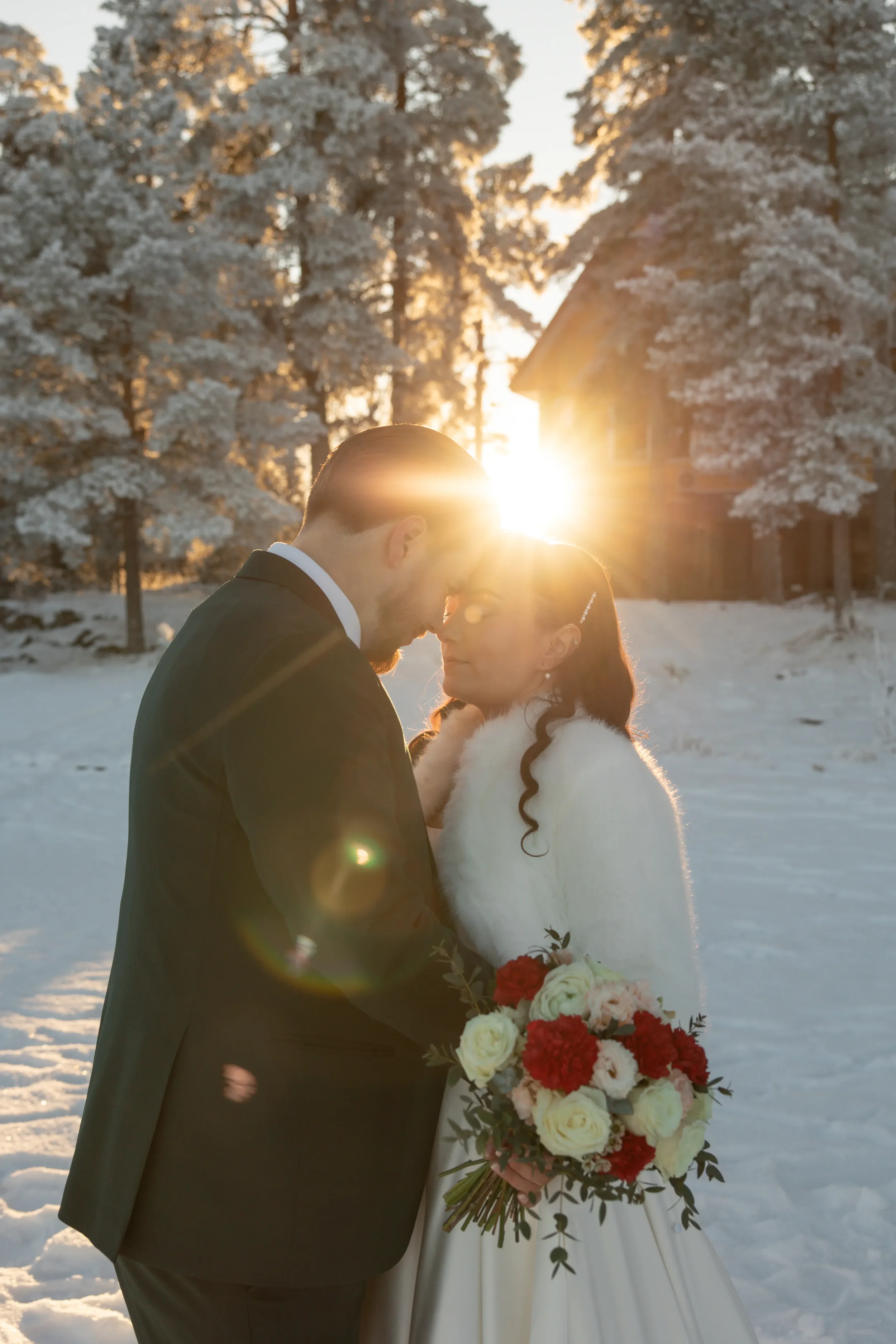 Hääpari otsat vastakkain talvimaisemassa auringonlaskussa, nainen pitelee kukkakimppua, taustalla lumiset puut ja talo. Hääkuvaaja Tampere.