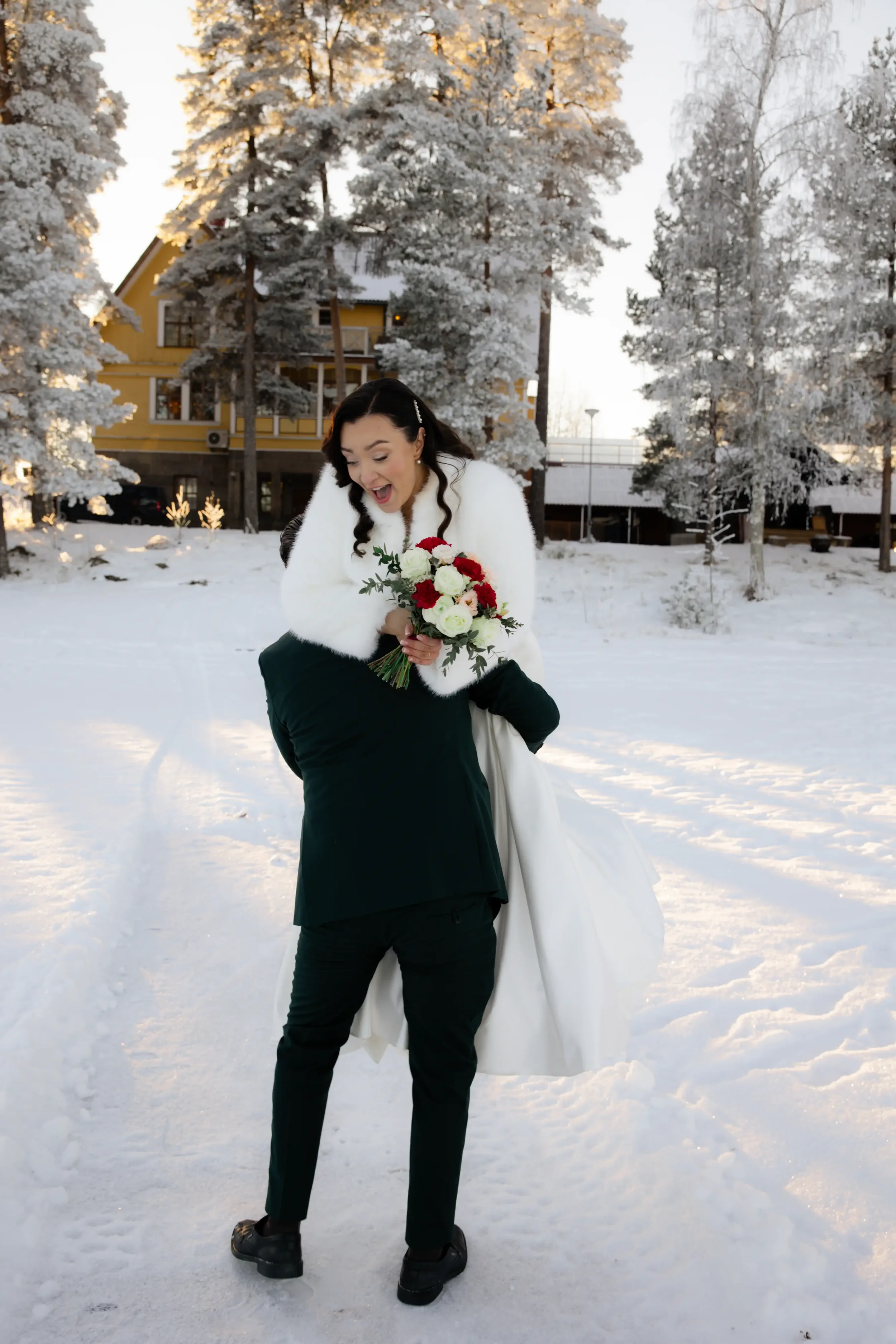 Hääpari juhlamielessä lumisessa maisemassa, morsian hyppää iloisesti suurta miestä päin ja pitää kukkakimppua, taustalla taloja ja lumiset puut. Hääkuvaaja Tampere.