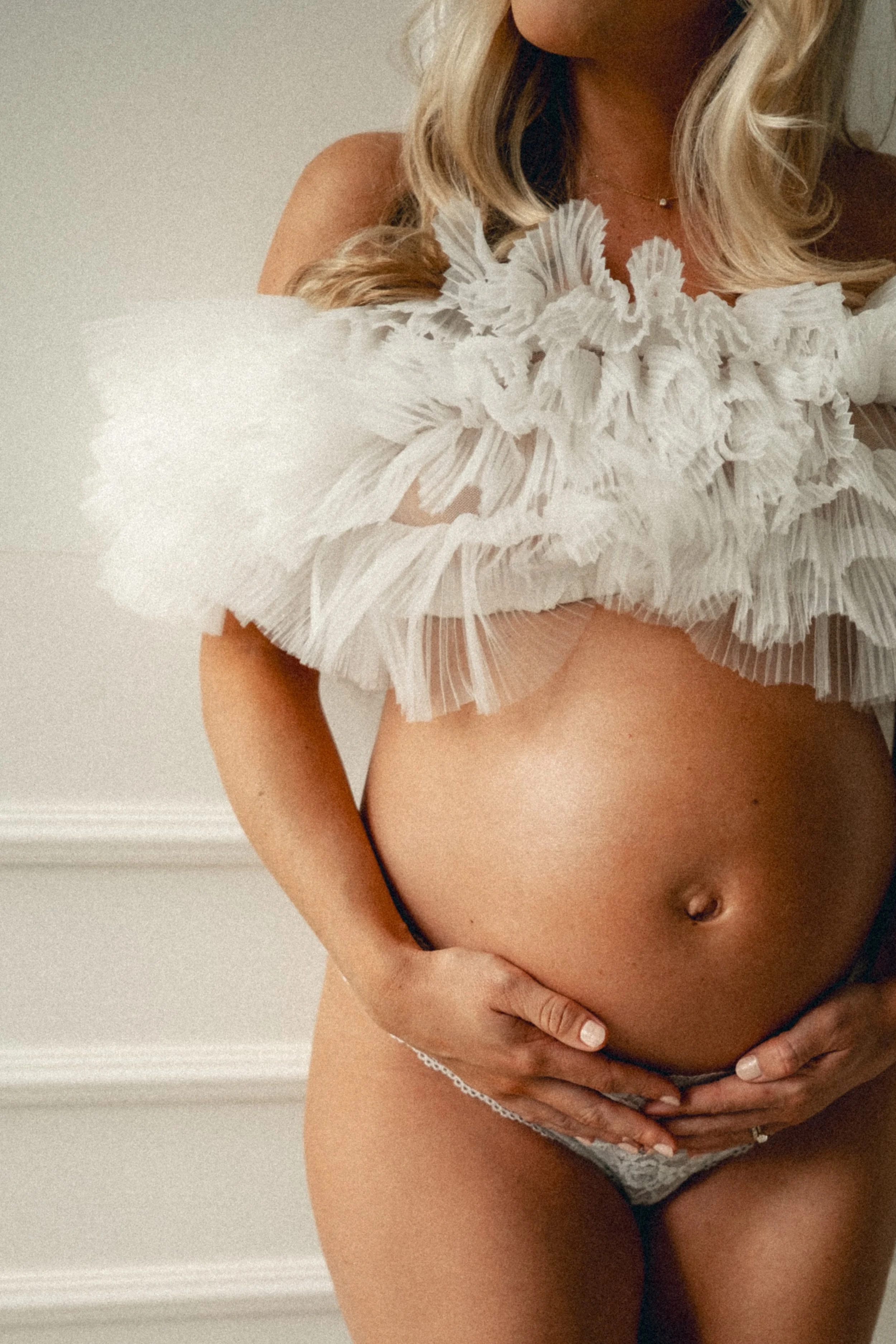 A pregnant woman with long blonde hair, wearing a ruffled white top and underwear, holding her belly with both hands.