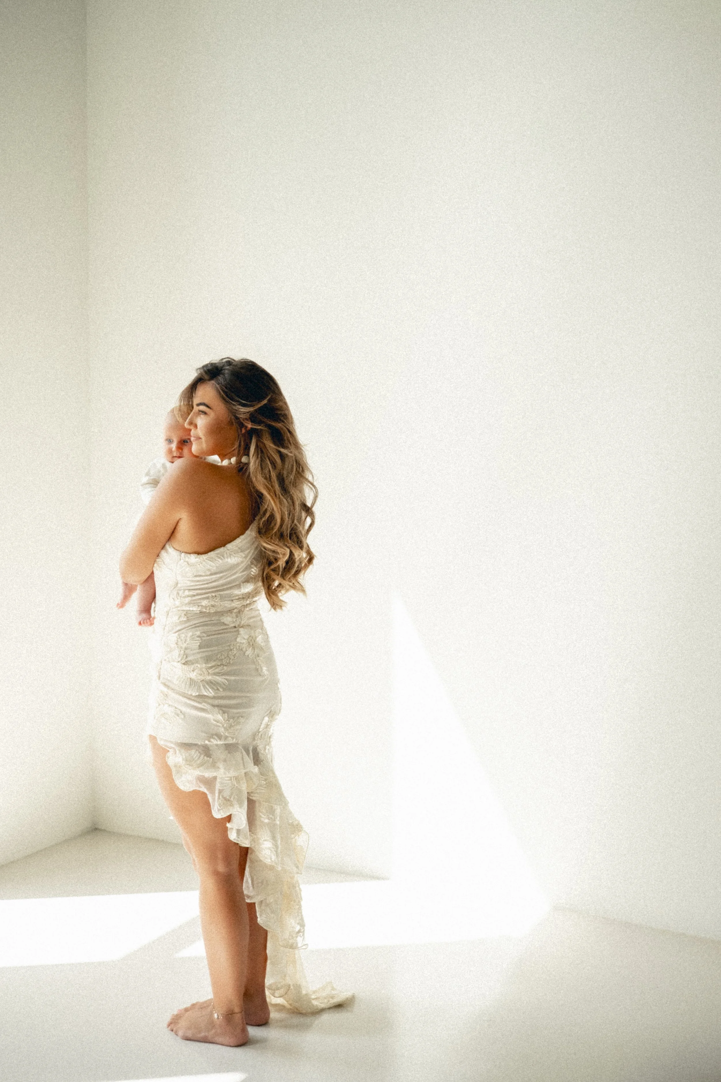 A woman in a ruffled white dress holding a baby in a white outfit, standing barefoot in a bright, minimal room with white walls and natural light.